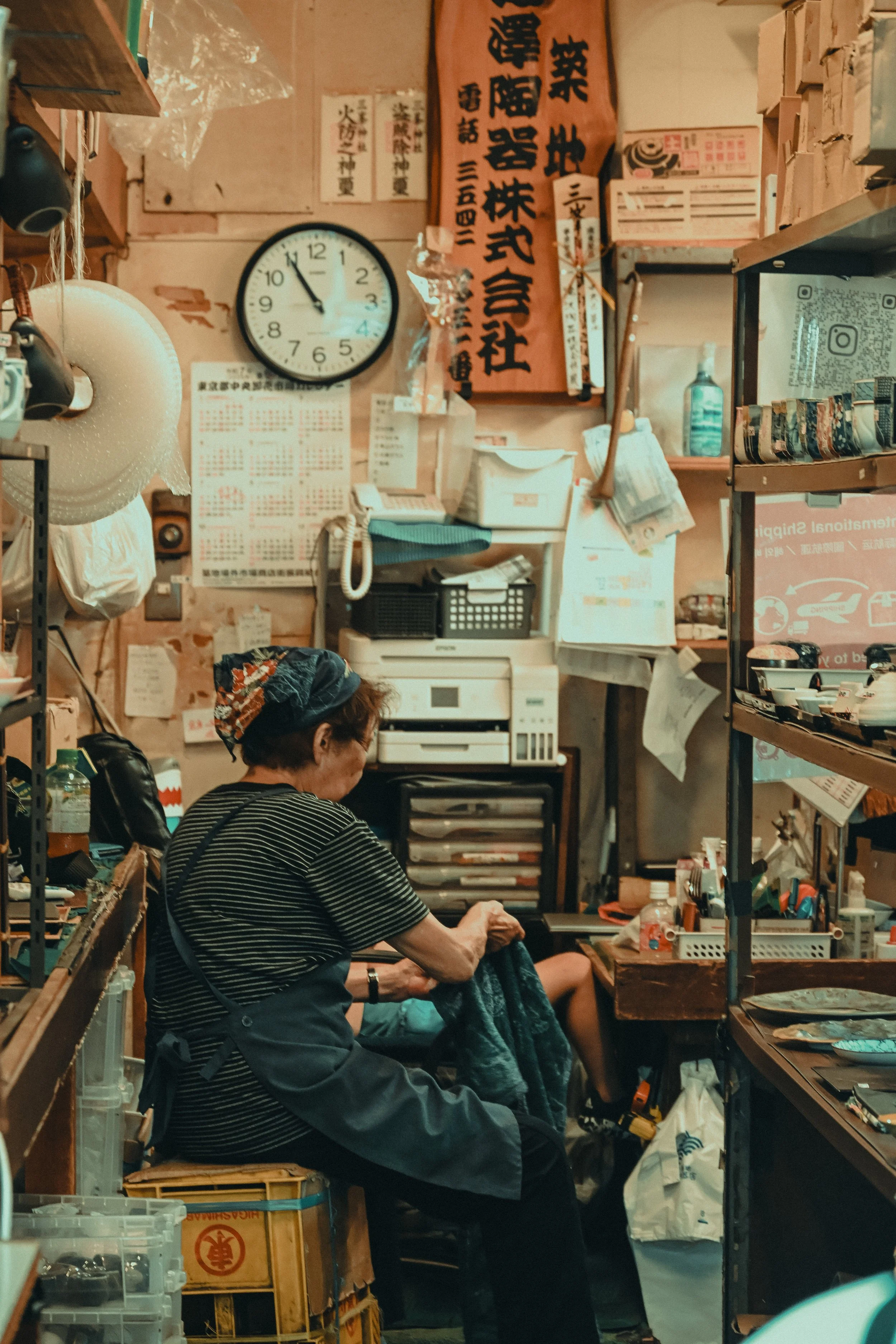 A woman with short hair and glasses, wearing a striped shirt and apron, sits at a cluttered workspace, working with dark fabric. The room is filled with boxes, shelves, and various objects including a clock showing 11:53, a large orange banner with J