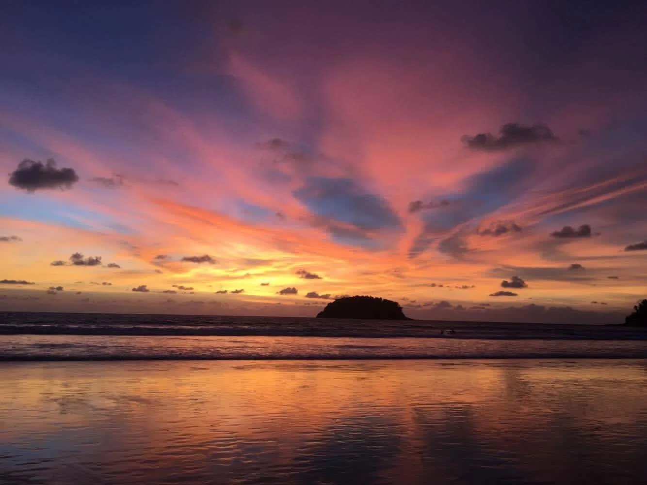 Sunset over the ocean with colorful sky and scattered clouds, with a silhouette of an island or landmass in the distance and its reflection on the water.