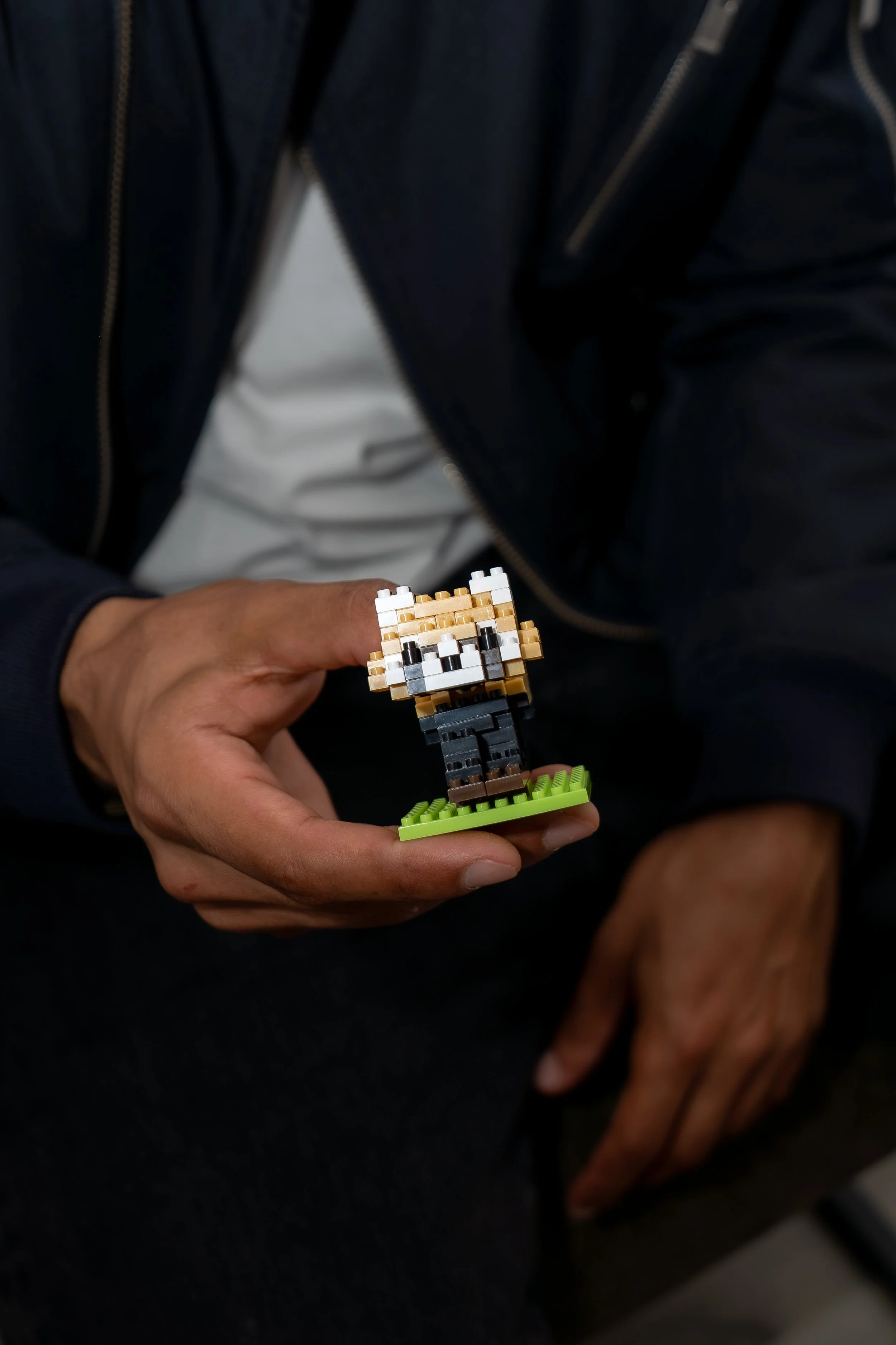 Close-up of a person's hand holding a small pixel art figurine of a dog, with a person's chest and arm in a dark jacket in the background.
