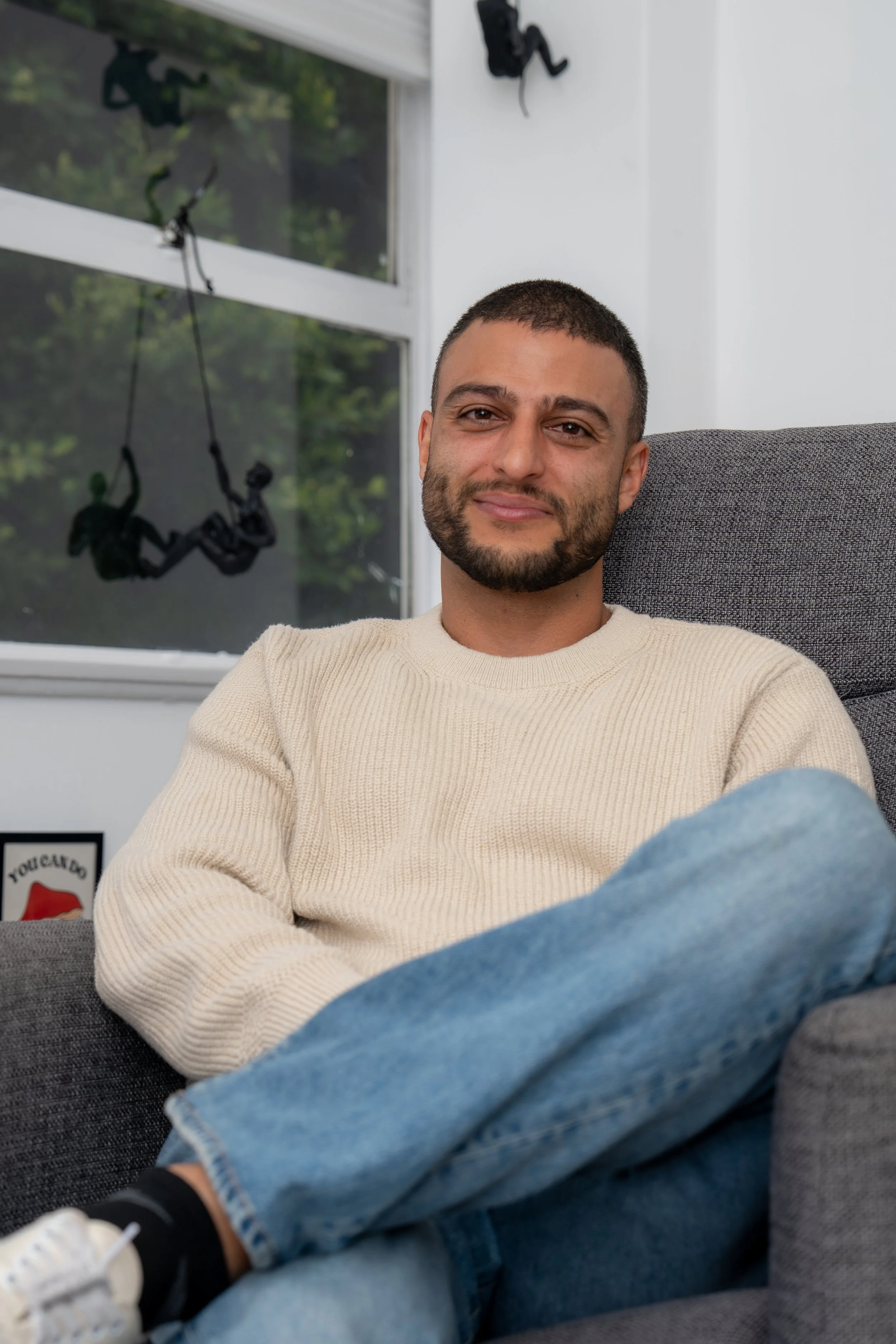 A man sitting on a gray couch in a room, wearing a beige sweater and blue jeans, with a window behind him showing a blurred outdoor scene and black wall decor hanging on the wall.
