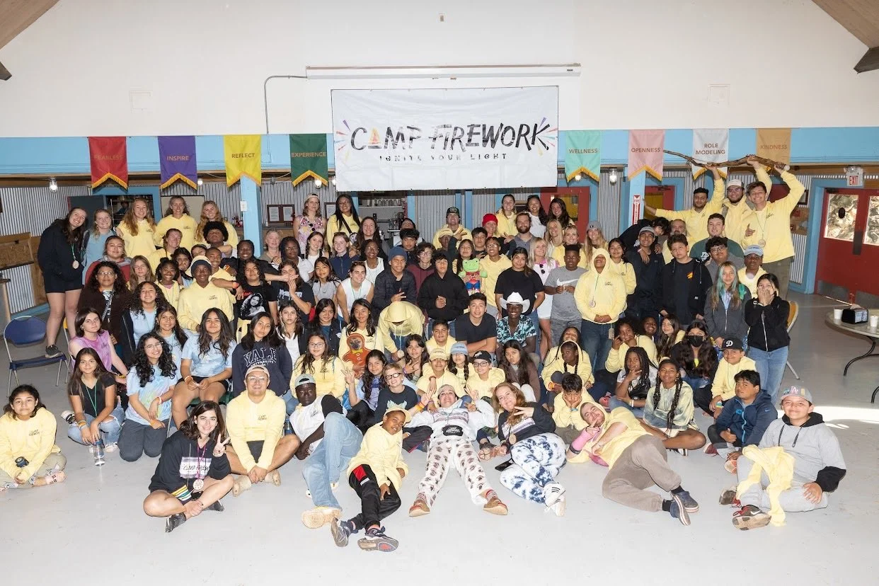 Group photo of children and teenagers at a campfire event in a large hall with colorful banners and a sign reading 'Camp Firework.' Many are smiling, some wearing yellow shirts, and some are sitting or standing in the front, middle, and back rows.