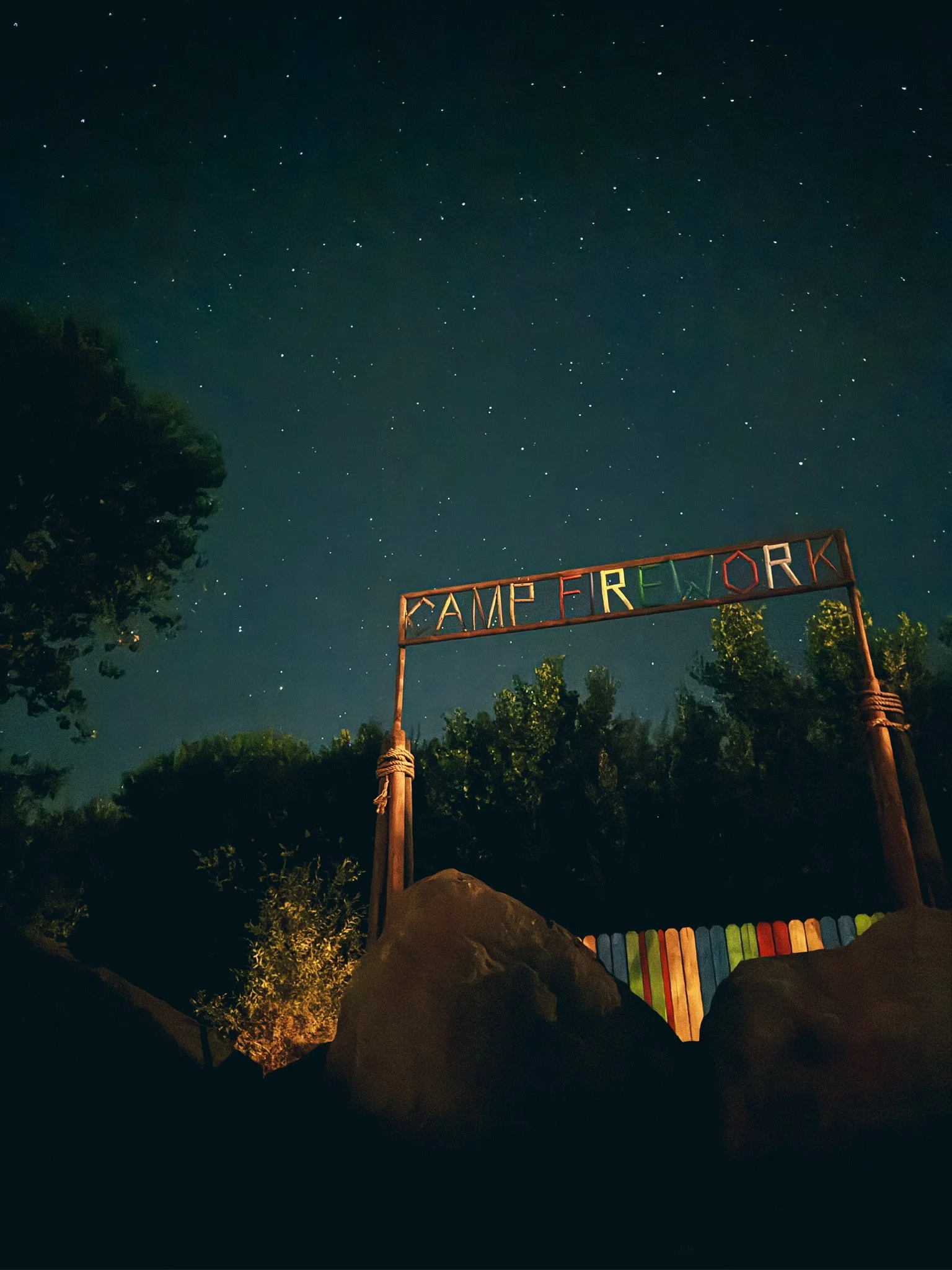 Night sky filled with stars with a campfire sign that reads 'CAMP FIREWORK' and a multicolored wooden fence in the foreground.