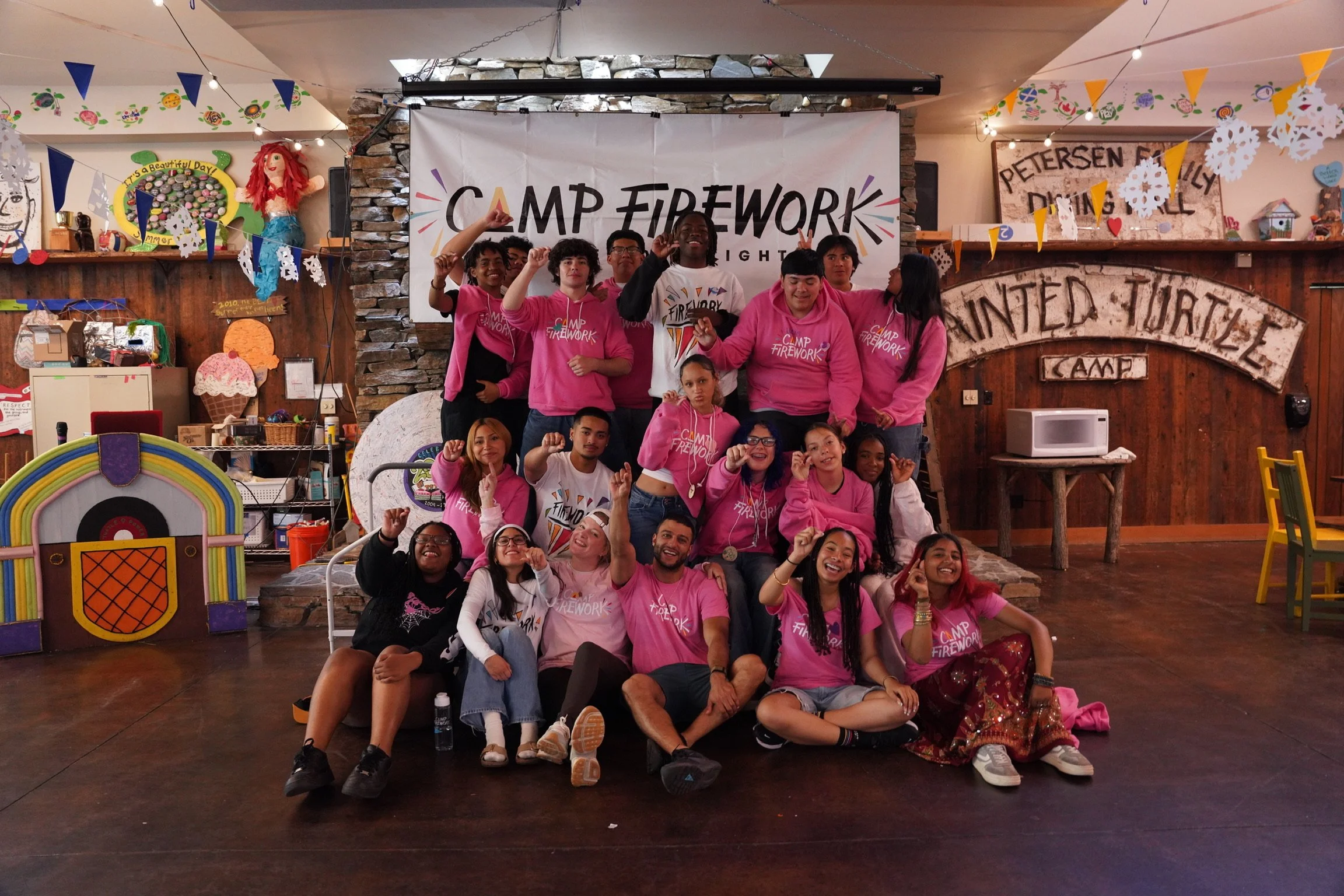 Large group of children and teenagers celebrating at a campfire themed event, wearing pink Camp Firework shirts, posing with raised fists and smiling, in a decorated indoor space with colorful banners and a banner reading 'Camp Firework' in the backg