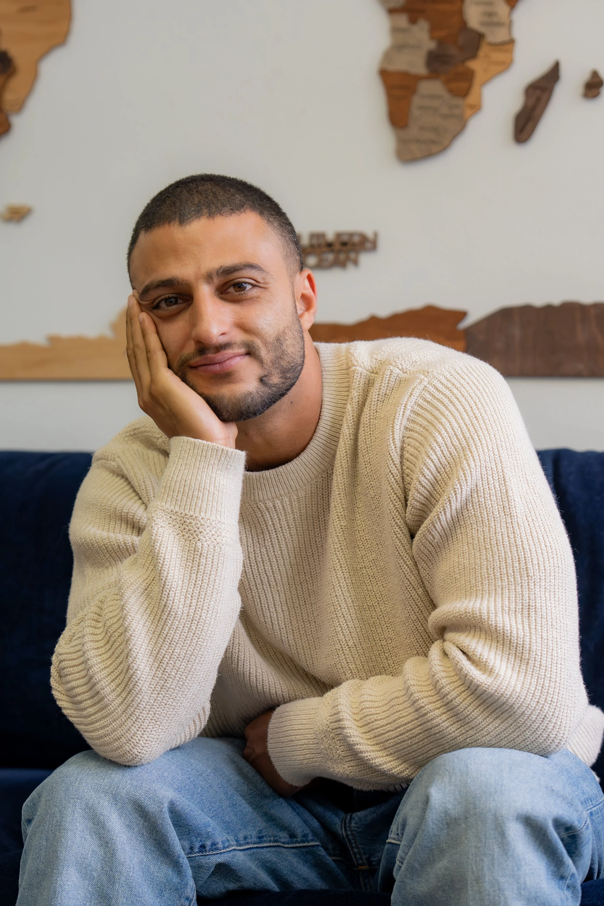 A young man with short dark hair and a beard, wearing a cream-colored sweater and blue jeans, sitting on a dark blue couch, resting his face on his hand, with a world map made of wooden pieces on the wall behind him.