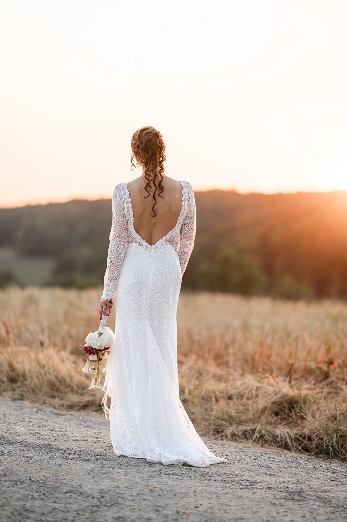 Rückansicht einer Braut im weißen Spitzenkleid mit Blumenstrauß bei Sonnenuntergang auf einem Feldweg