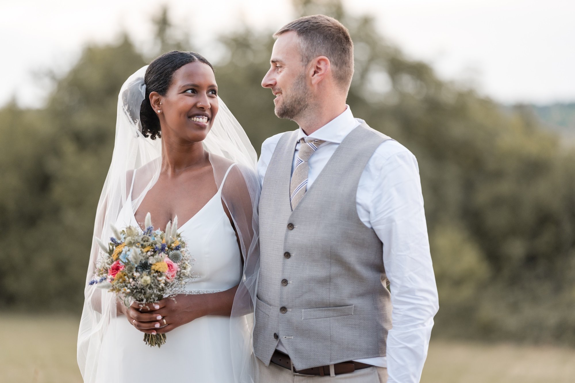 Ein Brautpaar bei einer Hochzeit im Freien. Die Braut trägt ein weißes Kleid und einen Schleier, hält einen Blumenstrauß, und lächelt, während sie den Bräutigam ansieht. Der Bräutigam trägt ein grau-weißen Anzug mit Krawatte. Sie schauen sich liebevoll an.