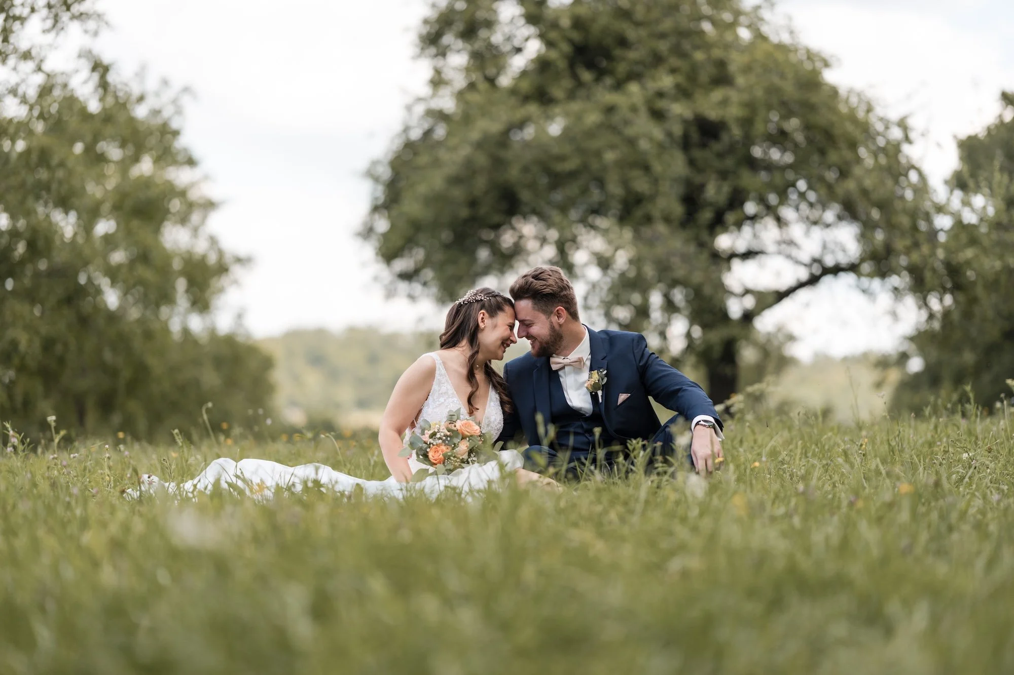 Ein junges Brautpaar sitzt lachend auf einer Wiese in Karlsruhe, umgeben von grünen Bäumen, bei einer Hochzeit im Freien.