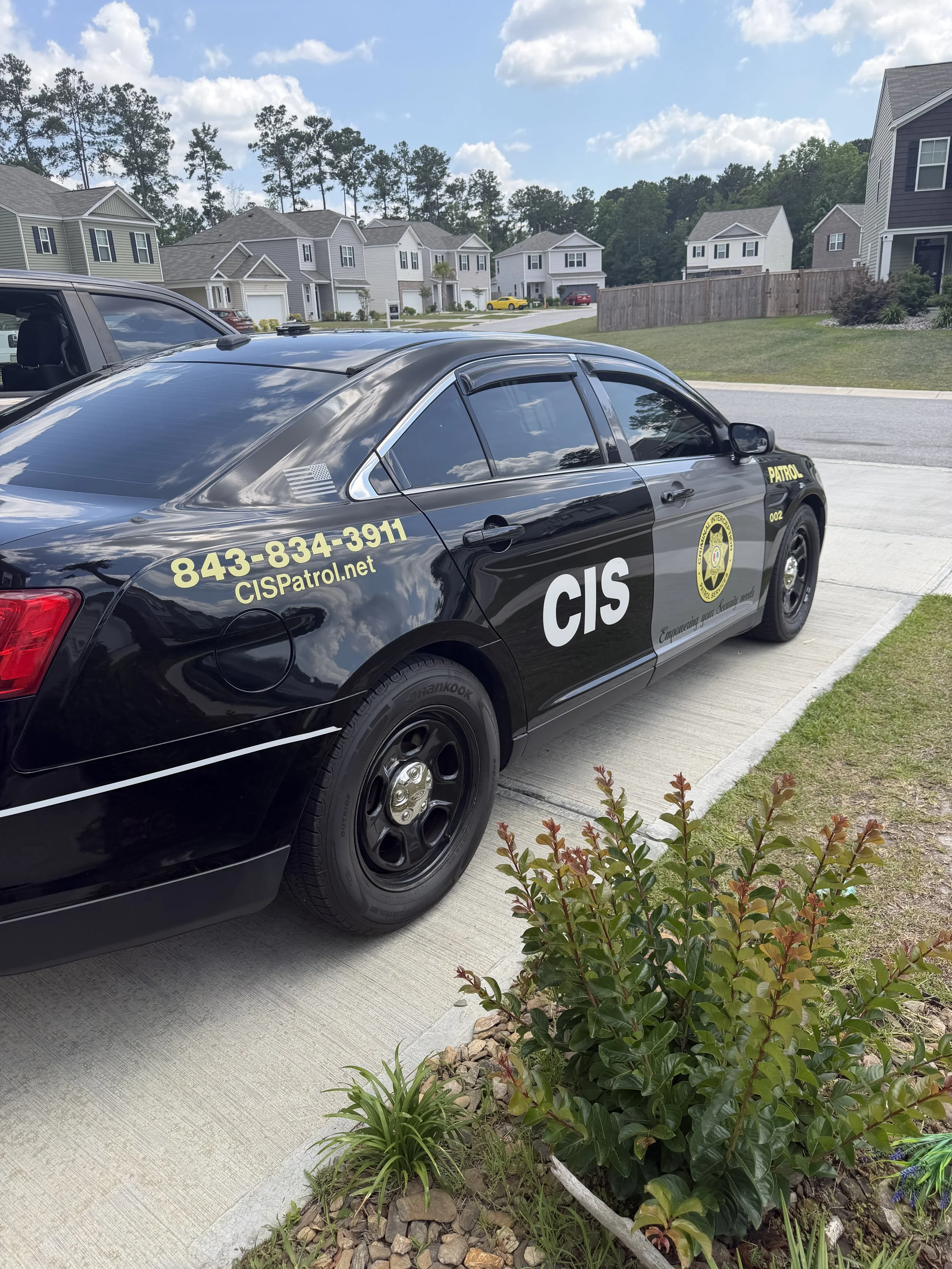 A black police patrol car parked on a residential street with houses and trees in the background. The car has CIS markings and contact information on the side.