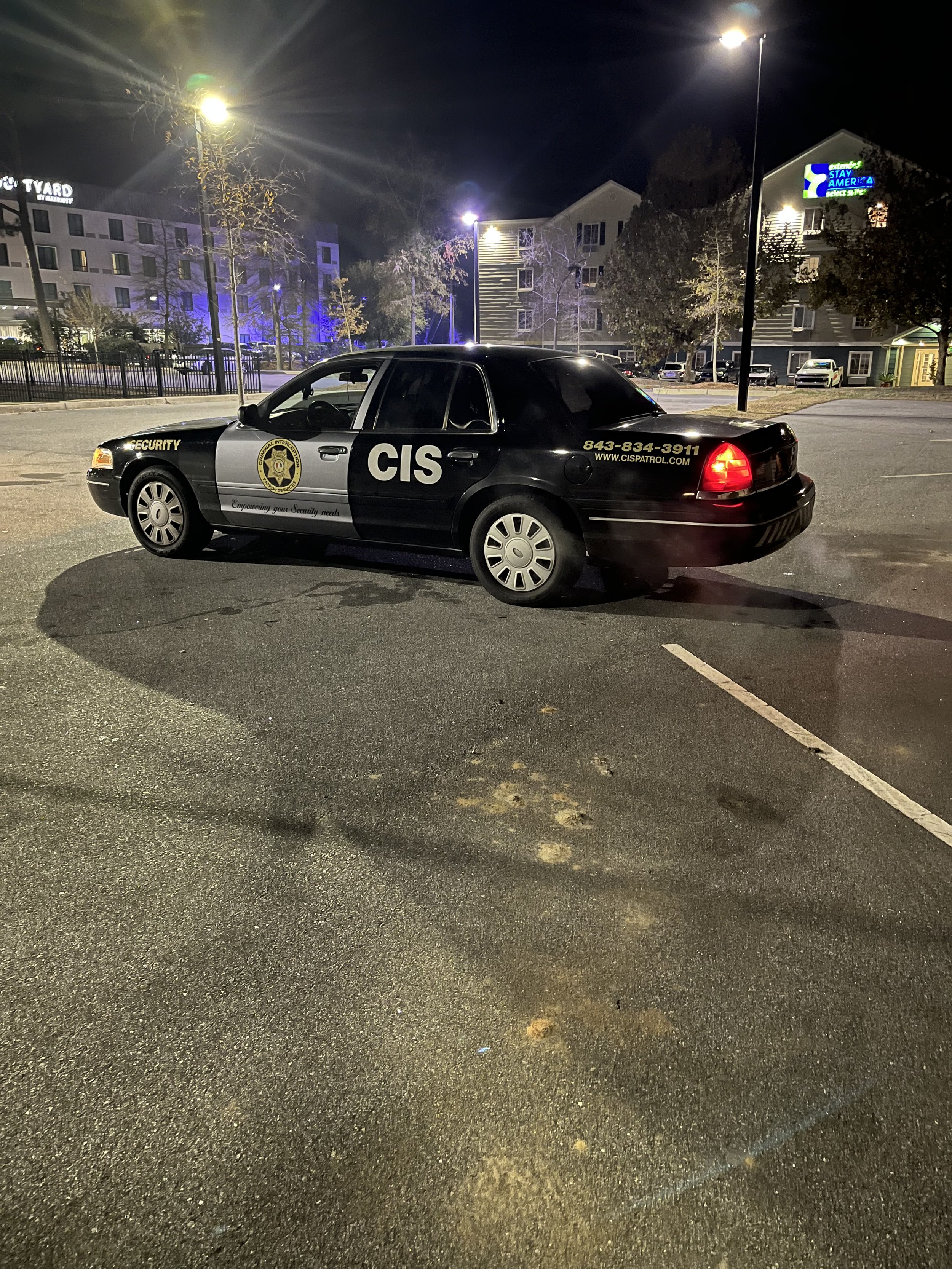 Nighttime parking lot with a police car marked CIS and Security parked in the foreground, buildings with lit windows and trees in the background.