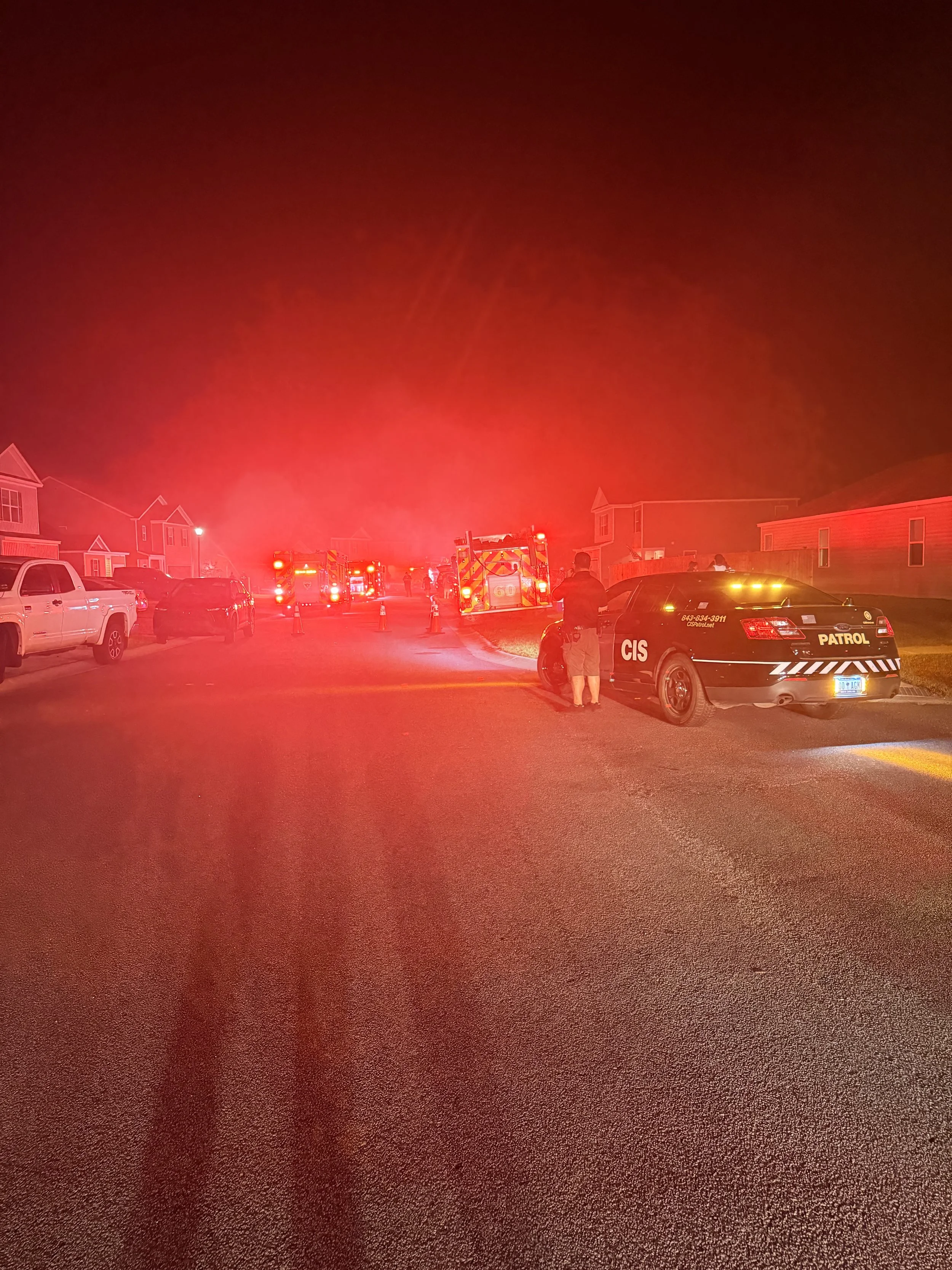 Nighttime scene of emergency vehicles and personnel responding to an incident in a residential neighborhood, with red emergency lights illuminating the scene.