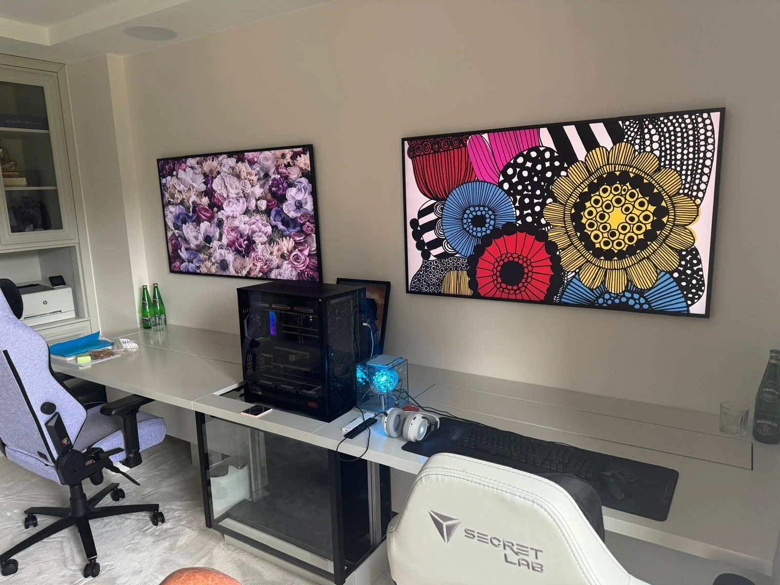 A modern home office with a white desk, a computer case, a headset, and a keyboard. Two large art prints hang on the wall; one features pink and purple flowers, the other has colorful abstract flower patterns. There is a white gaming chair with the '
