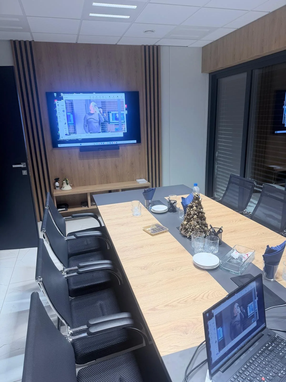 Conference room with a long wooden table, black chairs, a decorated Christmas tree, and a wall-mounted TV displaying part of the room. There are plates, glasses, and napkins on the table, along with a laptop and small decorative items.