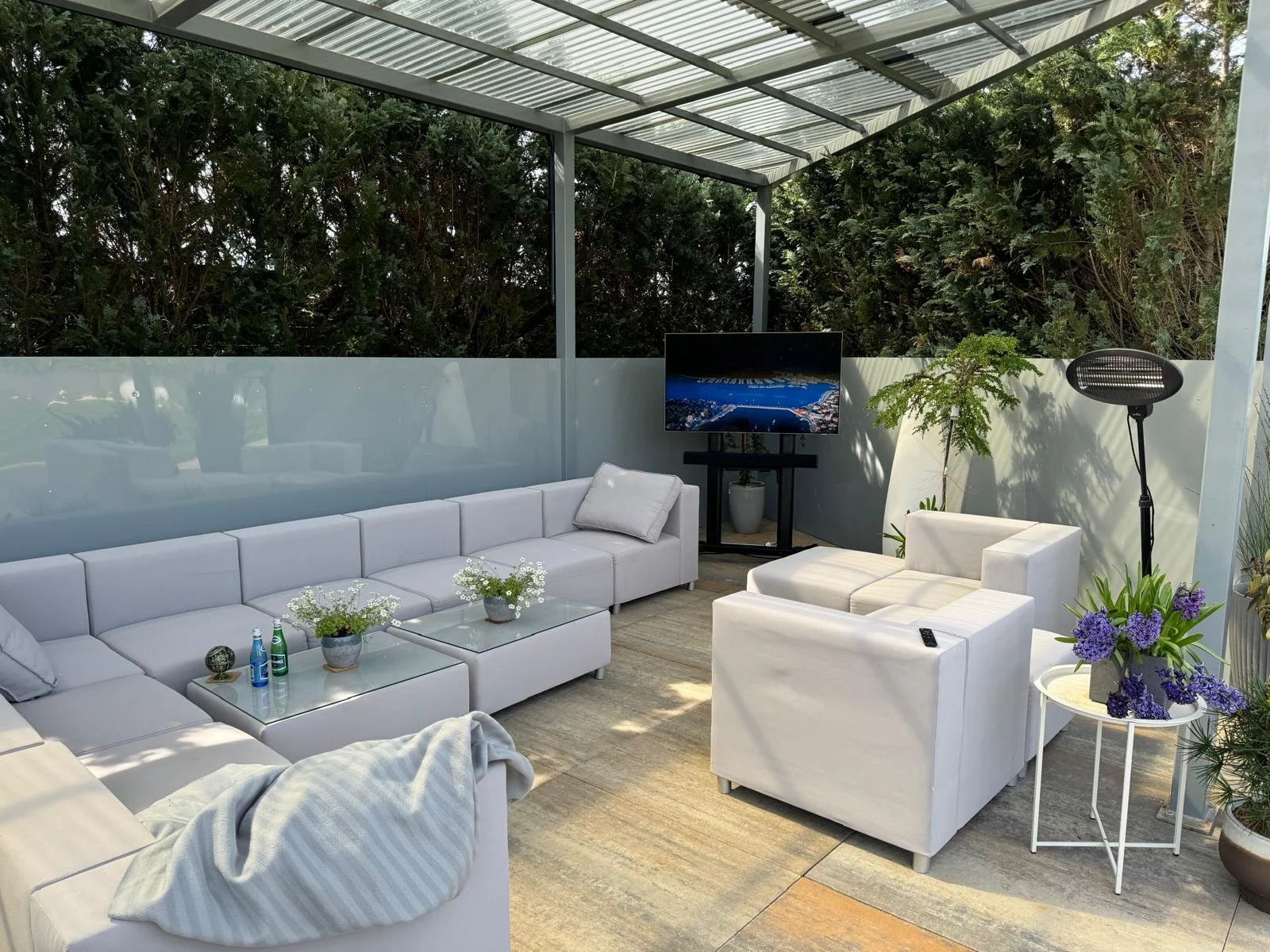 Outdoor patio with white sectional sofa, armchair, glass coffee table with plants, television, potted plants, and a standing lamp, surrounded by greenery.