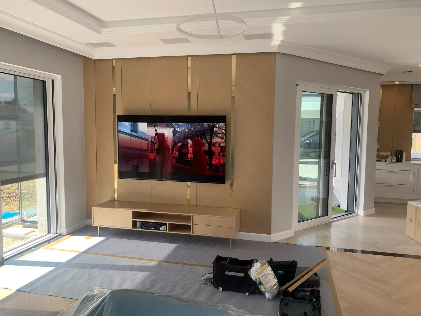 Modern living room with a mounted flat-screen TV on a beige decorative wall, a light wood media console, and large glass sliding doors leading outside. There are construction tools and materials on the floor.