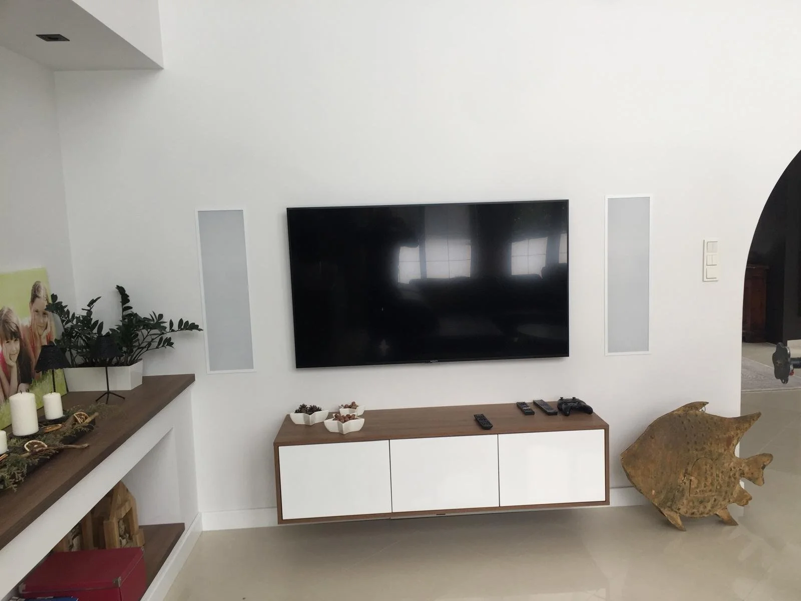 Living room with white walls, a mounted flat-screen TV, a white and wood TV stand, bowls of snacks, remote controls, a Game controller, artwork, candles, potted plant, and a wooden fish sculpture.