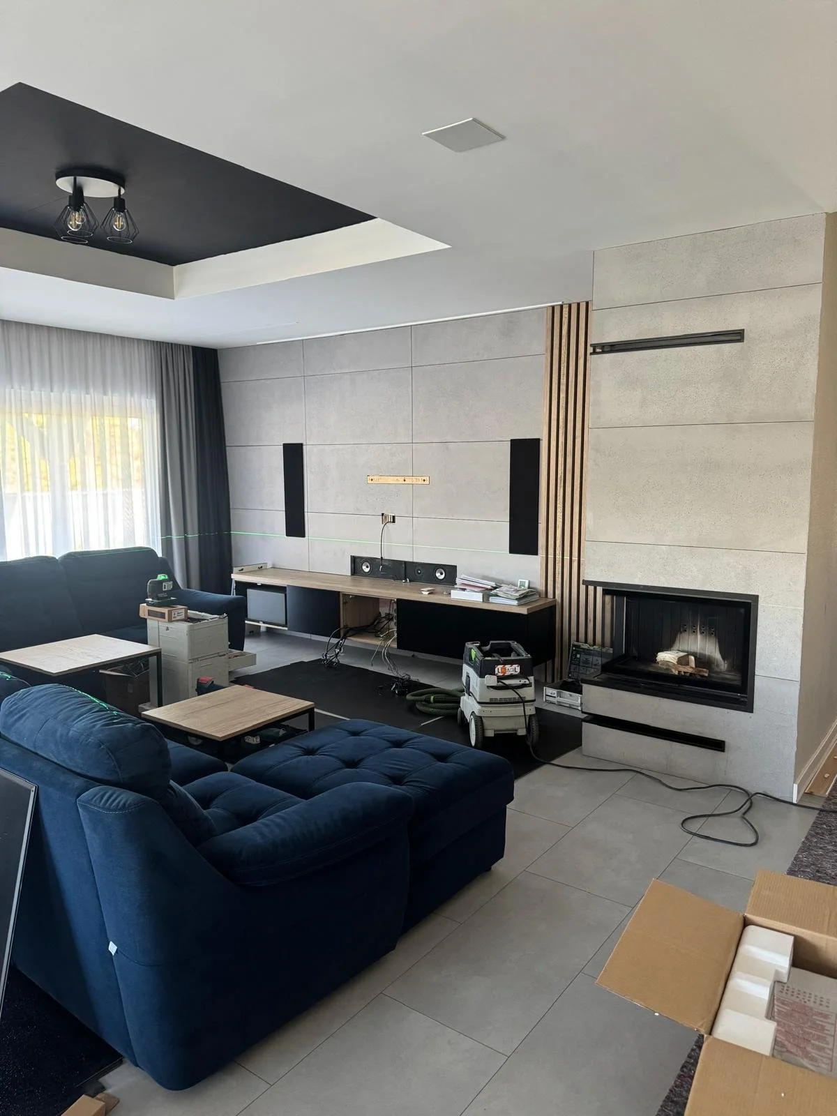 Living room with modern design, featuring a blue velvet sofa, a television wall, a fireplace, and living room furniture, with tools and equipment indicating ongoing setup or installation work.