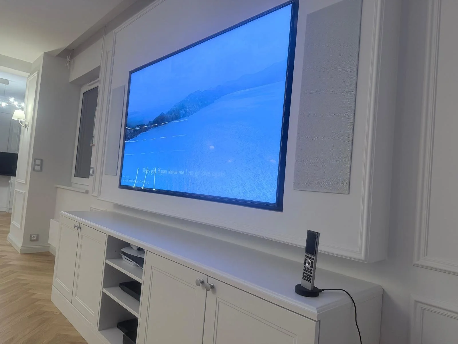 A living room with a large flat-screen TV mounted on a white wall, displaying a blue image of a body of water and mountains. Below the TV is a white cabinet with open shelves and a remote control on top.
