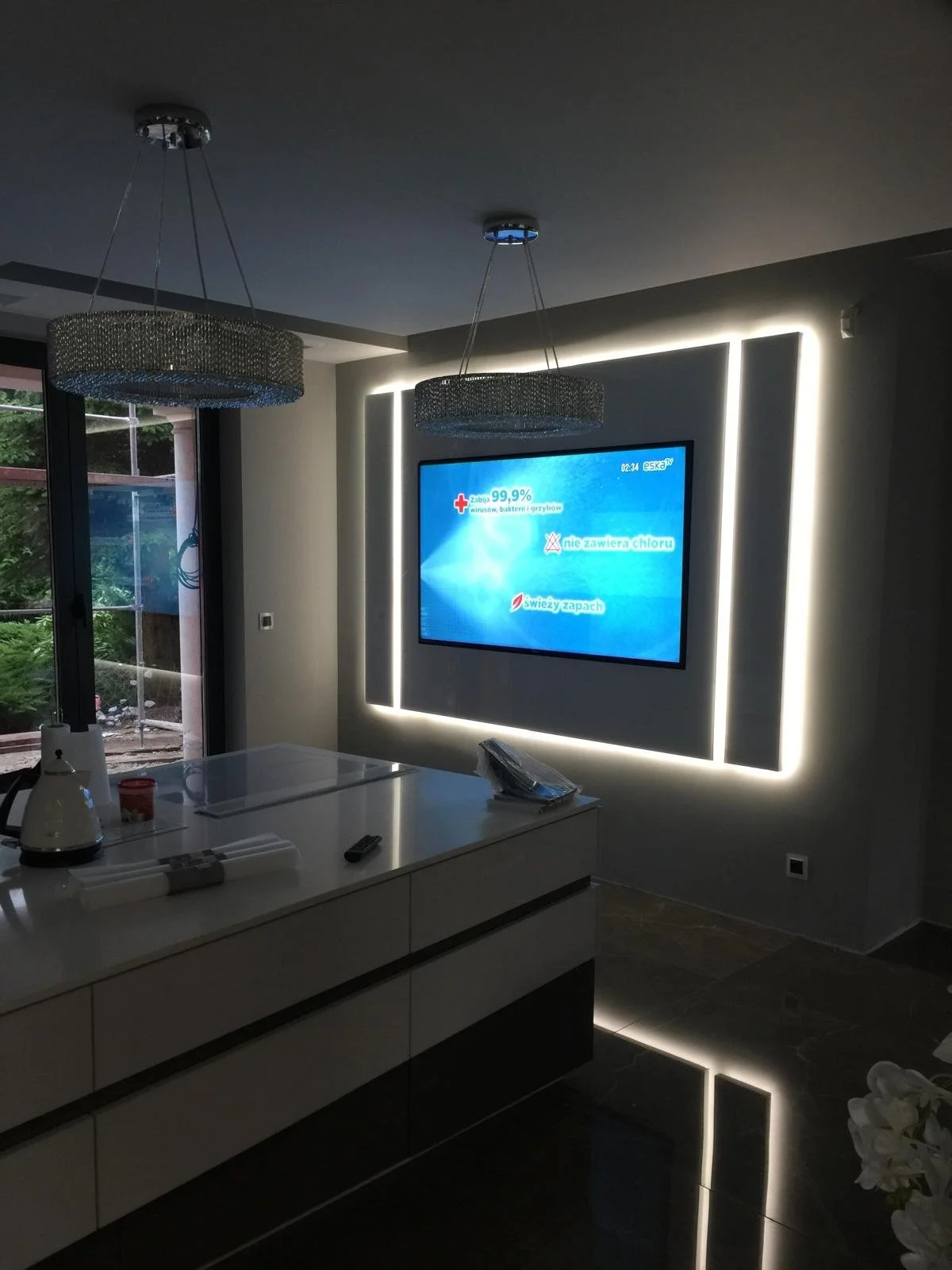 Modern living room with a large wall-mounted flat-screen TV surrounded by LED strip lighting, two decorative hanging light fixtures, a sliding glass door to the outside, and a white countertop with some kitchen appliances and a remote control on it.