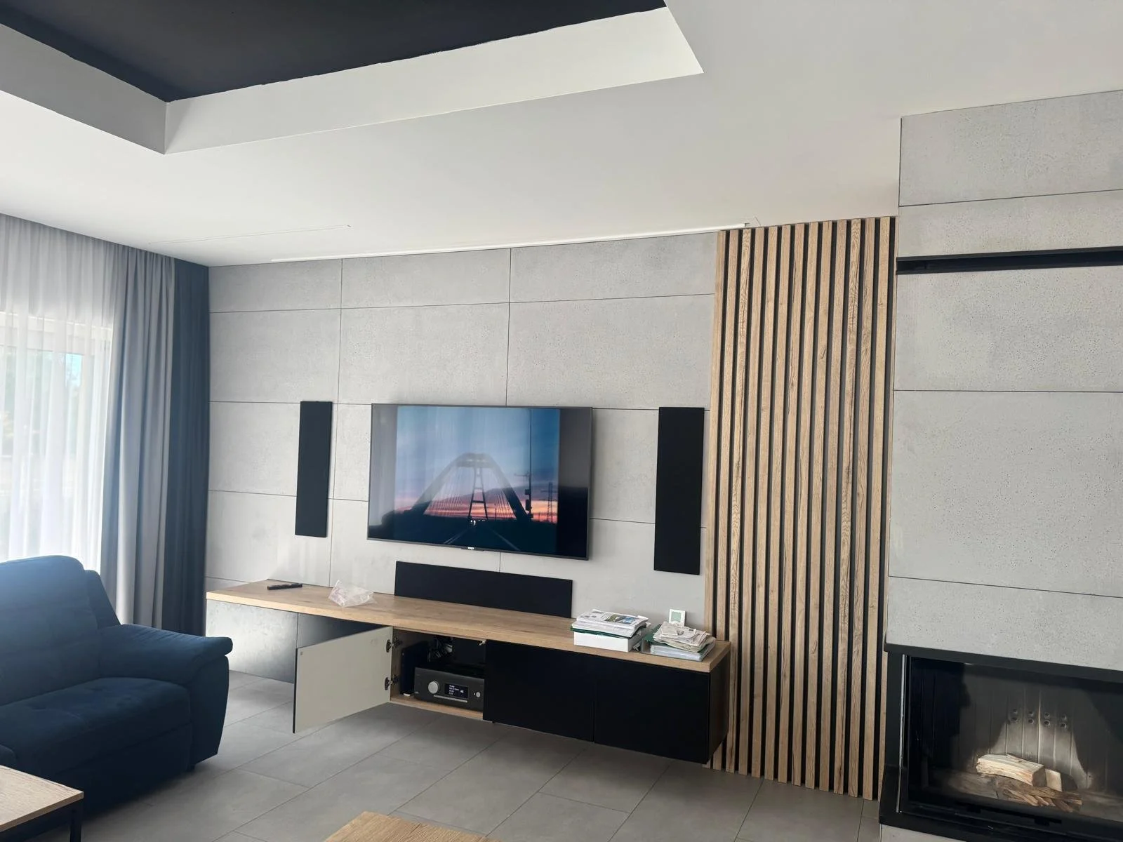 Modern living room wall with mounted flat-screen TV displaying a bridge at sunset, black soundbar and speakers, wooden panel design, gray tiled walls, and a small fireplace.