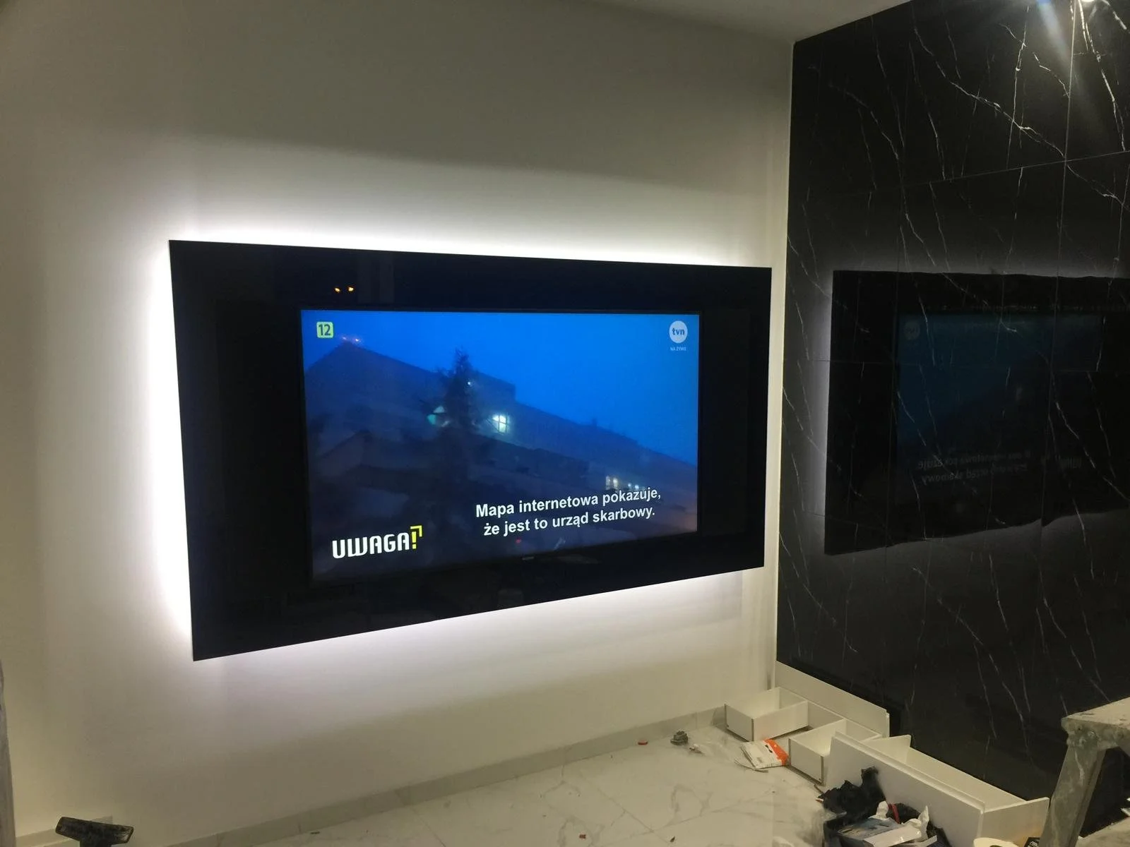 A television screen installed on a wall displaying a warning message in Polish, with some construction tools and debris on the floor below.