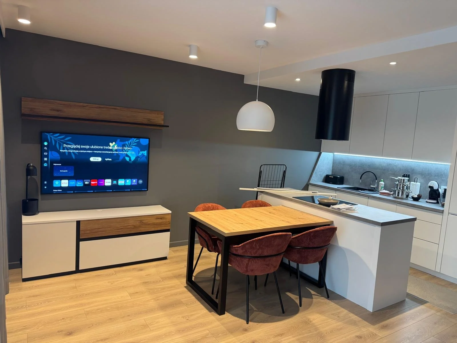 Modern open-plan kitchen with a wooden dining table, four red upholstered chairs, a grey wall with a mounted television, a black range hood, and minimalistic white cabinets and countertops.