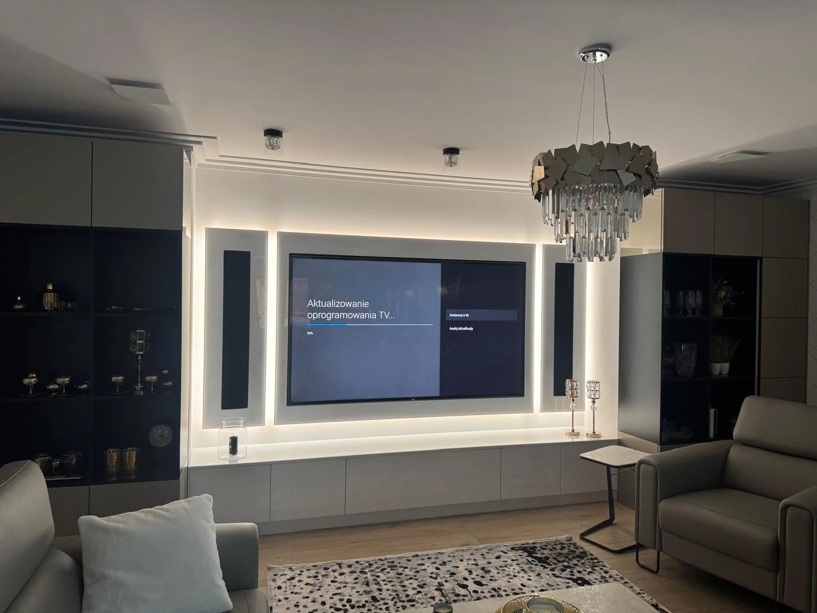 Modern living room with a large flat-screen TV displaying a TV software update message, surrounded by built-in shelves with decor, a chandelier hanging from the ceiling, and seating including a beige sofa with white pillows and a matching armchair.
