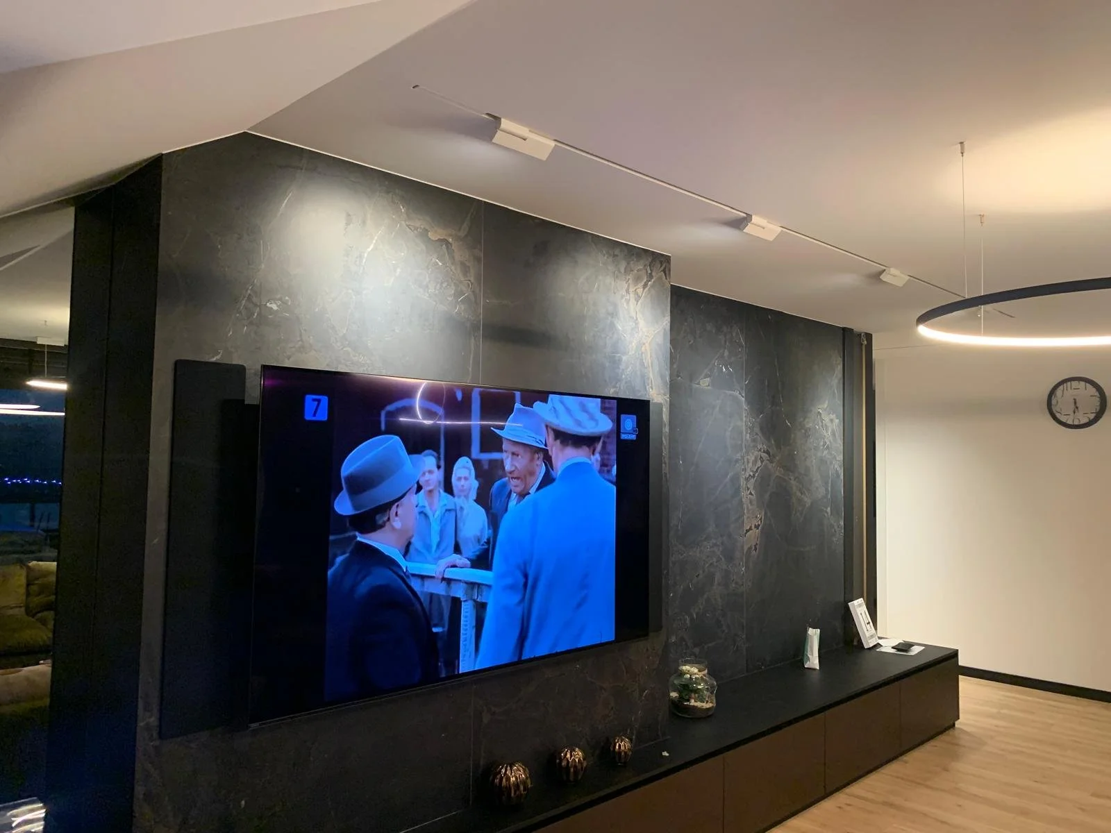Living room with a large flat-screen TV on a black marble wall, displaying a scene with people in vintage attire. To the right, a modern circular ceiling light and a wall clock are visible. A black console below the TV holds small decorative objects 