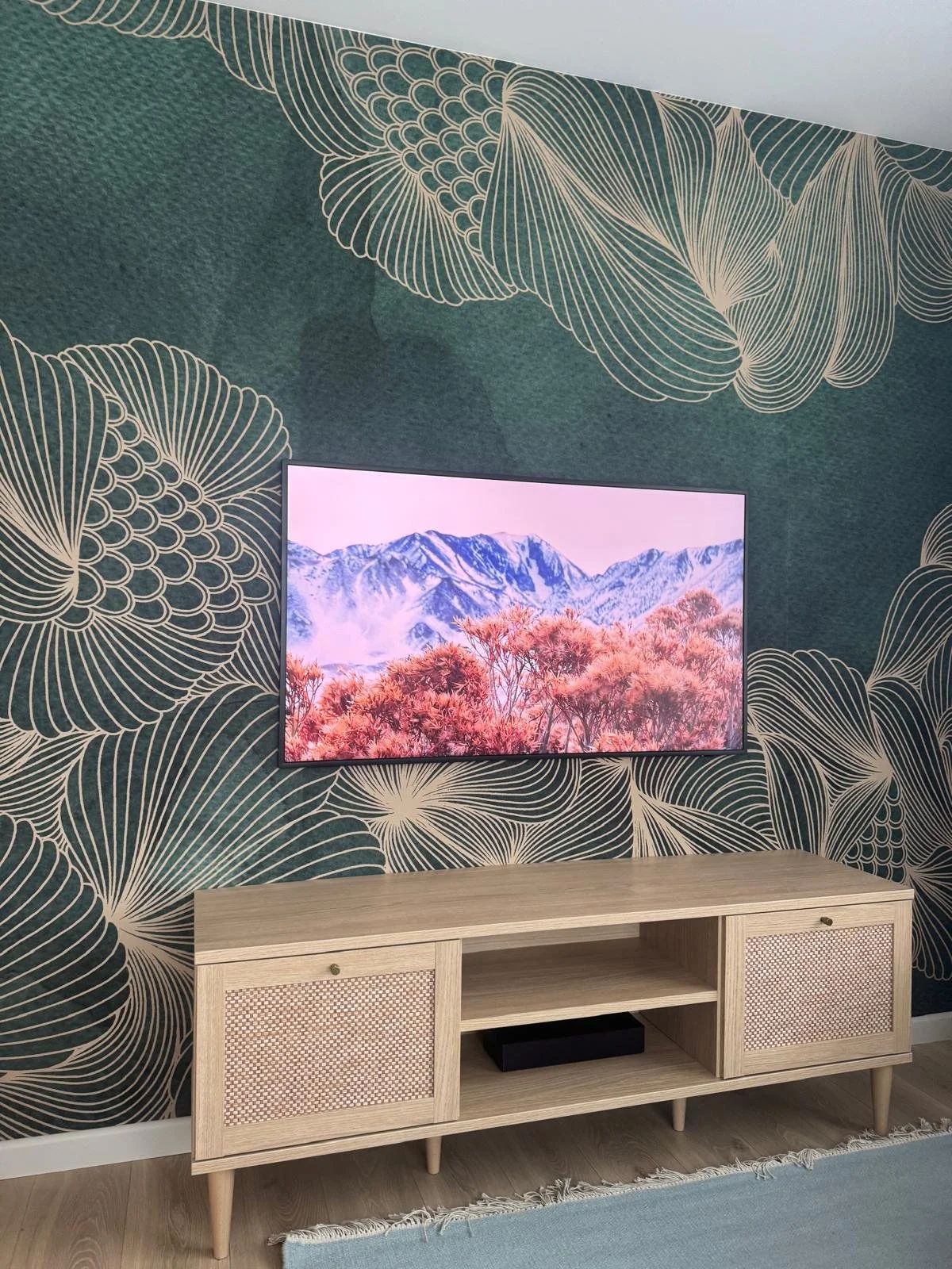 Living room with a wall-mounted flat screen TV showing a mountain landscape with pink trees and snow-capped peaks. The wall has dark teal wallpaper with large, flowing, white botanical patterns. Below the TV is a light wooden console table with two s