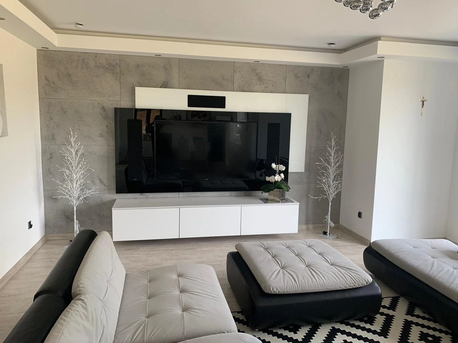Living room with beige and black furniture, a large flat-screen TV mounted on a textured gray wall, white sideboard, two white decorative trees, a potted plant, black and white geometric rug, and ceiling light fixtures.
