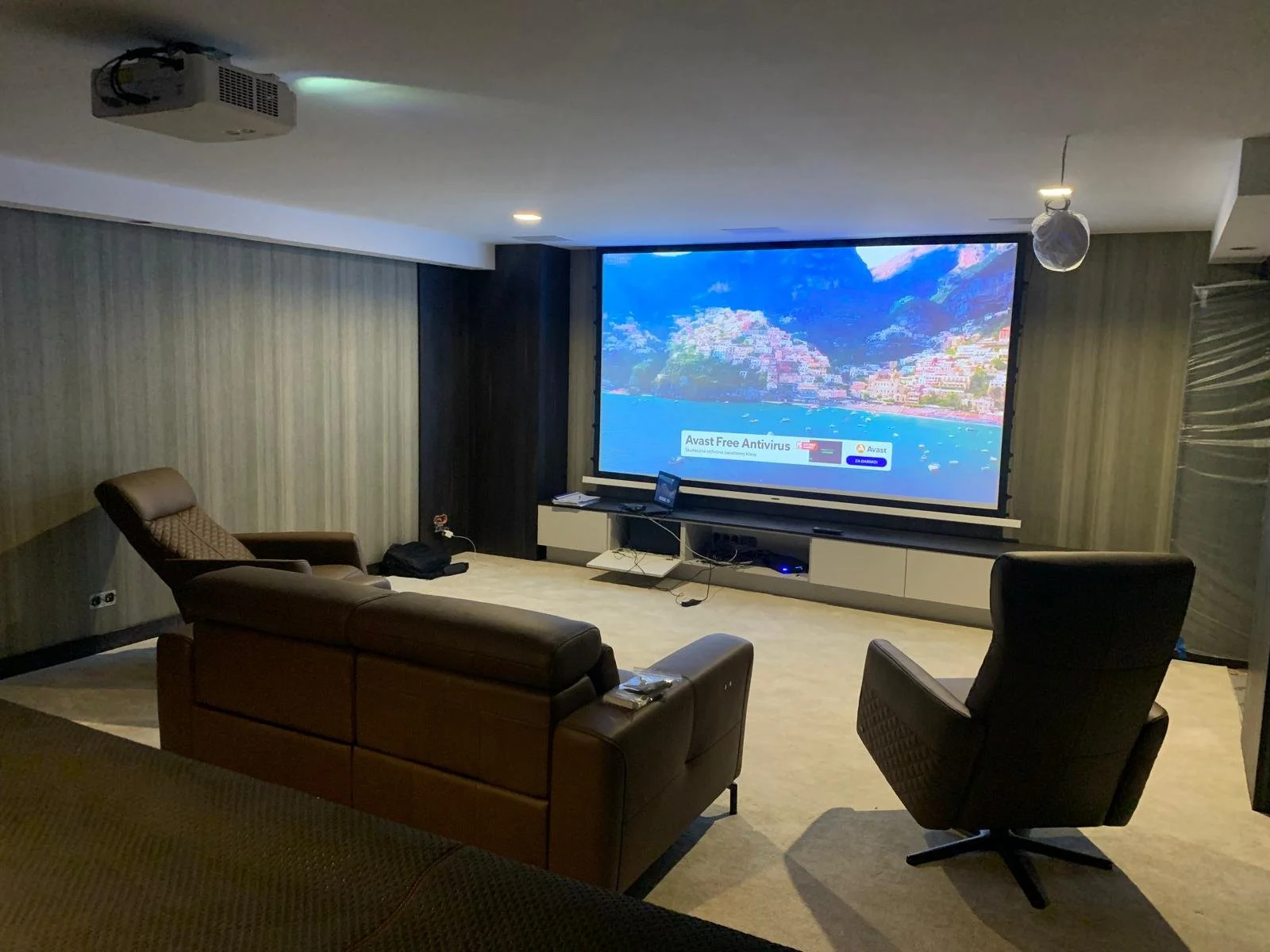 A modern home theater with a large screen, some reclining and standard chairs, and a projector mounted on the ceiling.