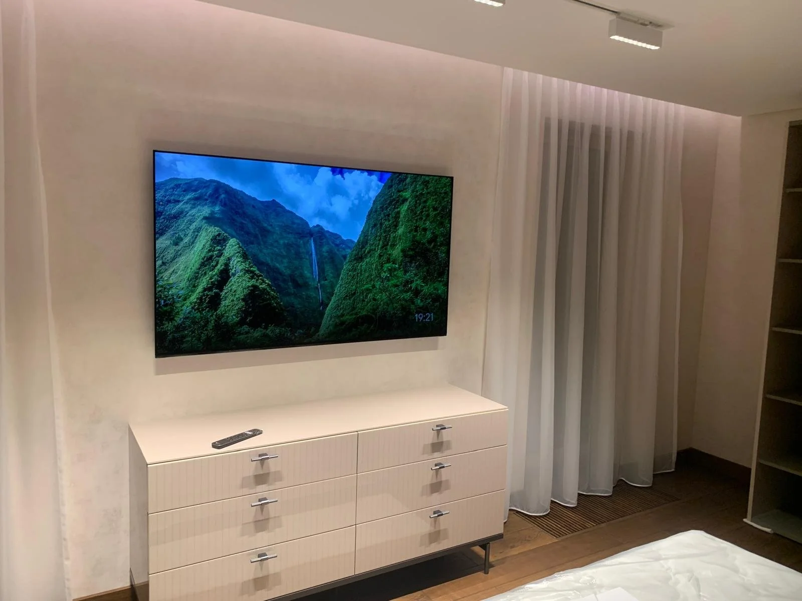 Living room with a wall-mounted flat-screen TV displaying a mountain landscape, white dresser beneath the TV with a remote control on top, sheer white curtains to the right, and part of a bed in the foreground.