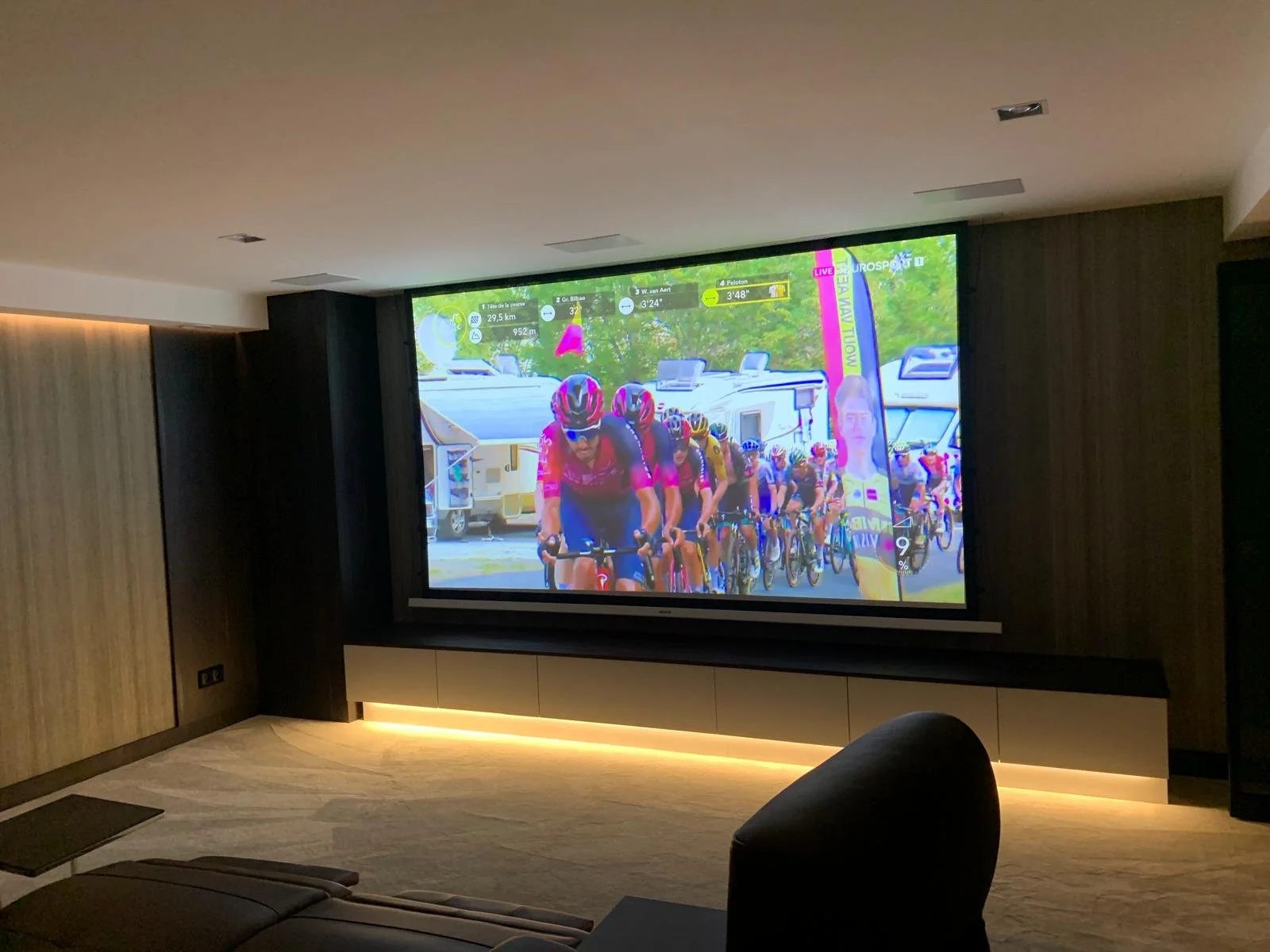 Indoor room with a large screen displaying a cycling race. The screen shows a group of cyclists riding outdoors with trees and campers in the background.