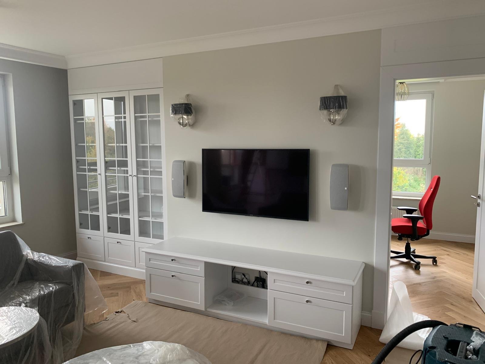 Living room with a wall-mounted flat-screen TV, white cabinet, and two wall-mounted speakers. A doorway leads to an office with a red chair and a window showing trees outside.