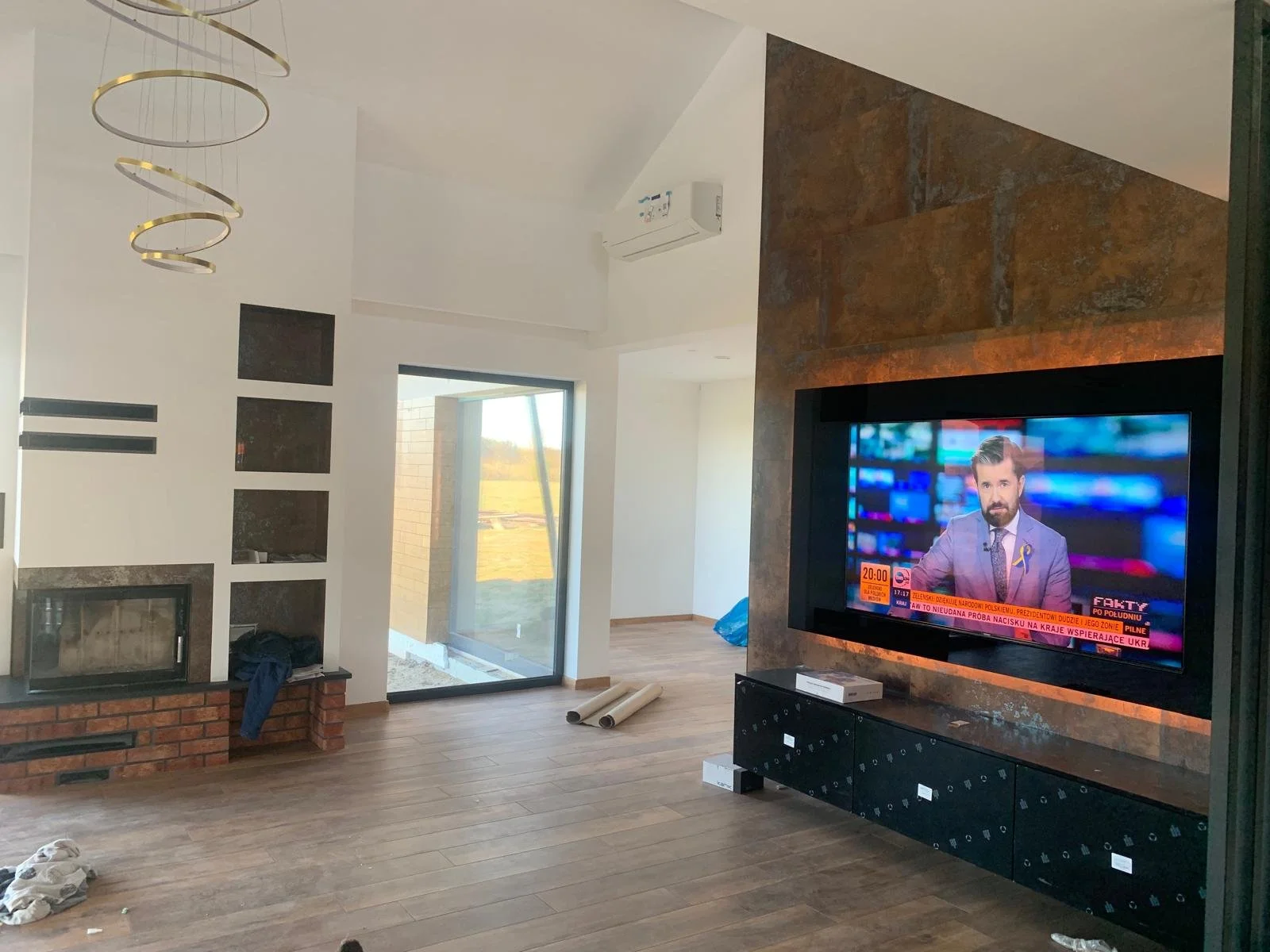 Modern living room with a mounted flat-screen television on a brick and wood wall, a fireplace with brick surround below a white wall with built-in shelves, a sliding glass door leading outside, a tall white air conditioning unit, wood flooring, and 