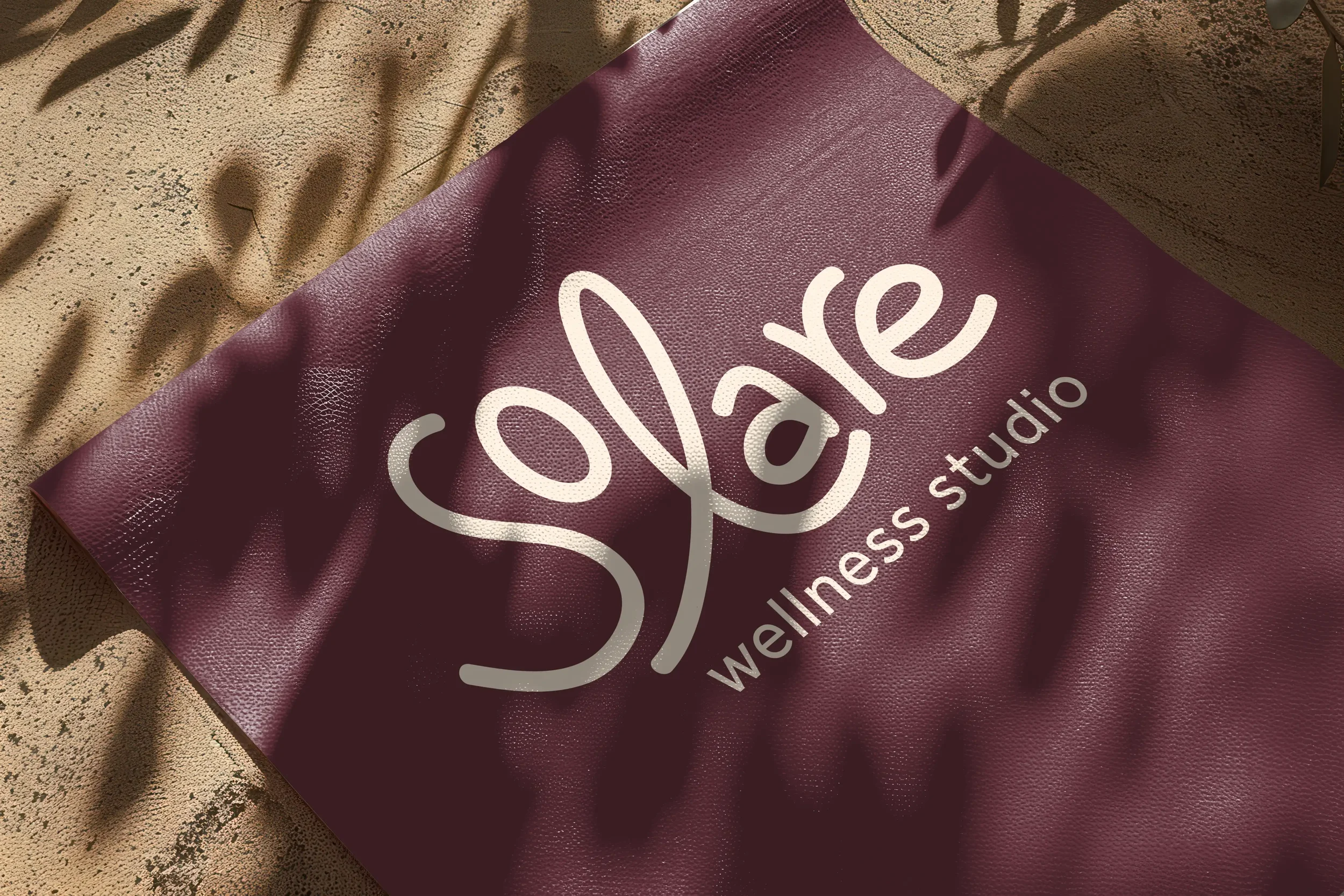 A burgundy rectangular signage board with the word 'Yale' written in large white cursive letters, and partially visible smaller text below. The board is placed on a textured beige surface and has shadows of leaves cast across it.