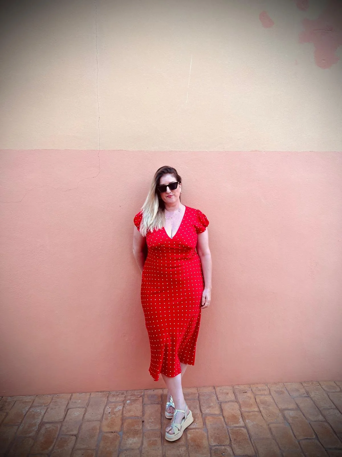 A woman in a red polka dot dress and sunglasses standing against a pink and beige wall.