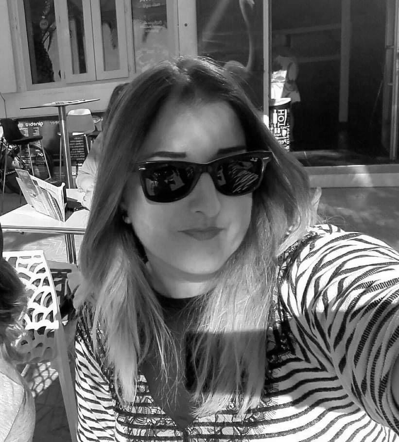 A woman with long hair wearing sunglasses and a striped shirt, taking a selfie outside in a sunny environment.