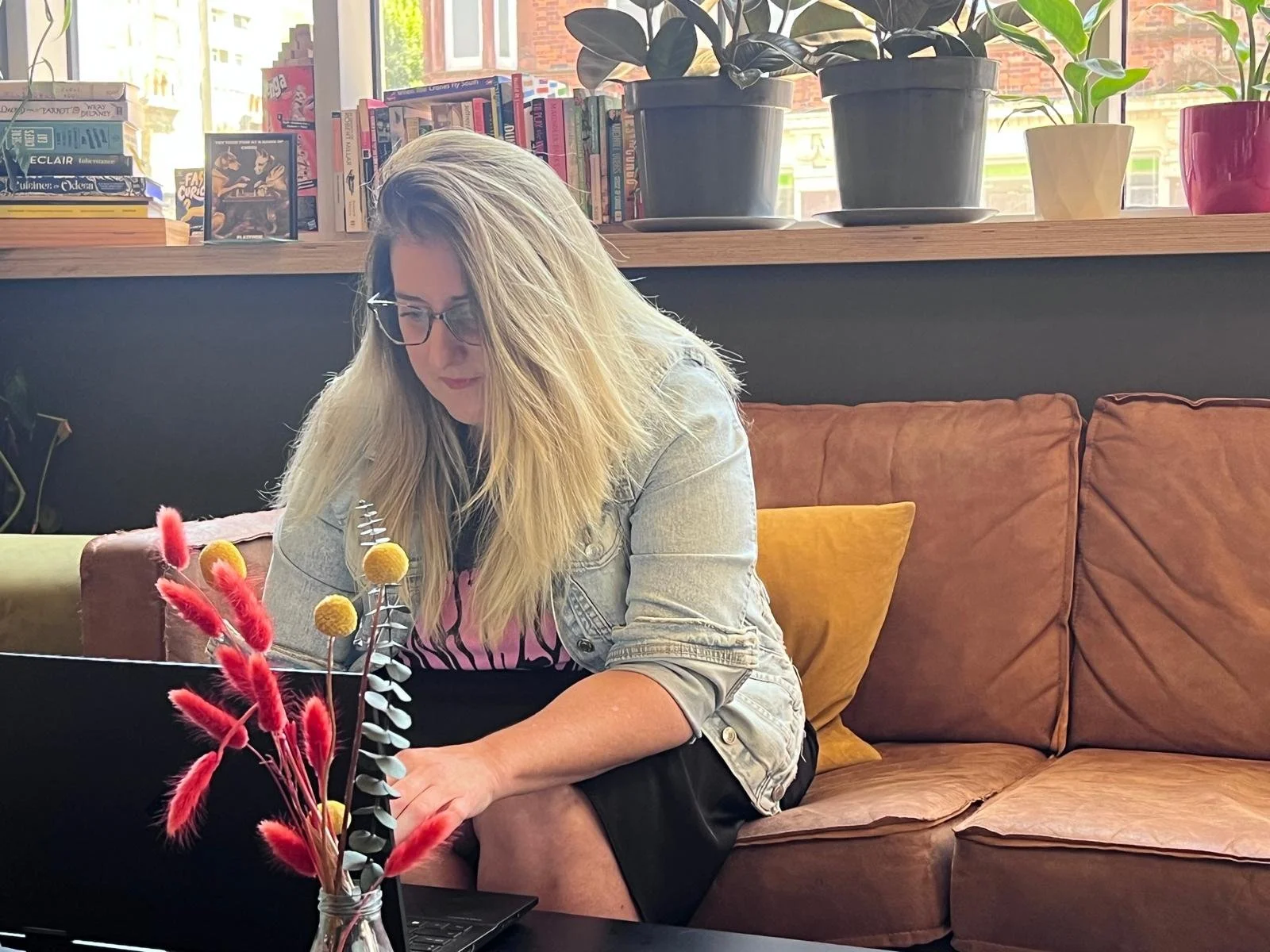 A woman with long blonde hair and glasses sitting on a brown leather couch, working on a laptop in a cozy room filled with books and potted plants.