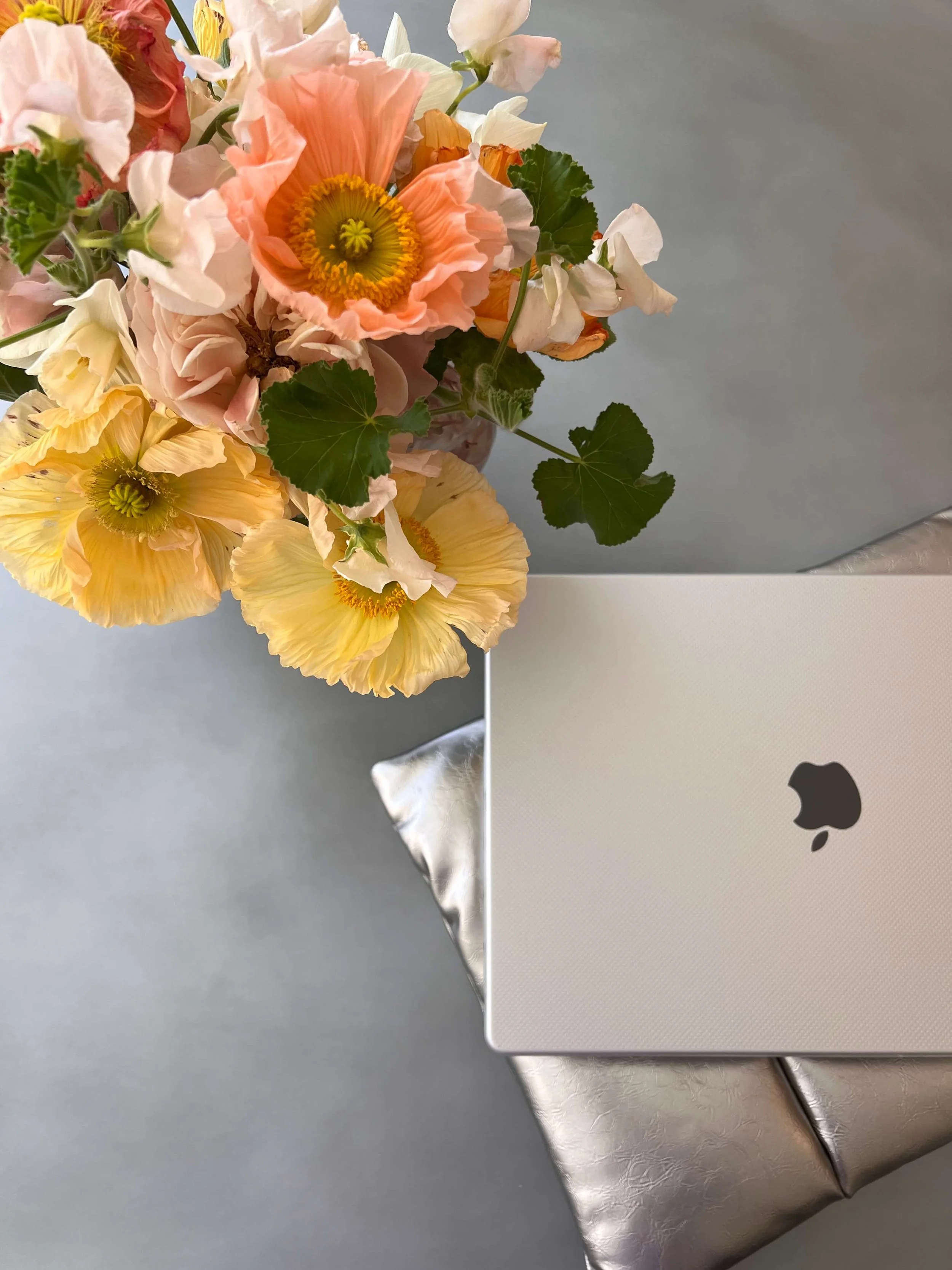 A bouquet of pink, white, and yellow flowers with green leaves on a table next to a silver laptop.