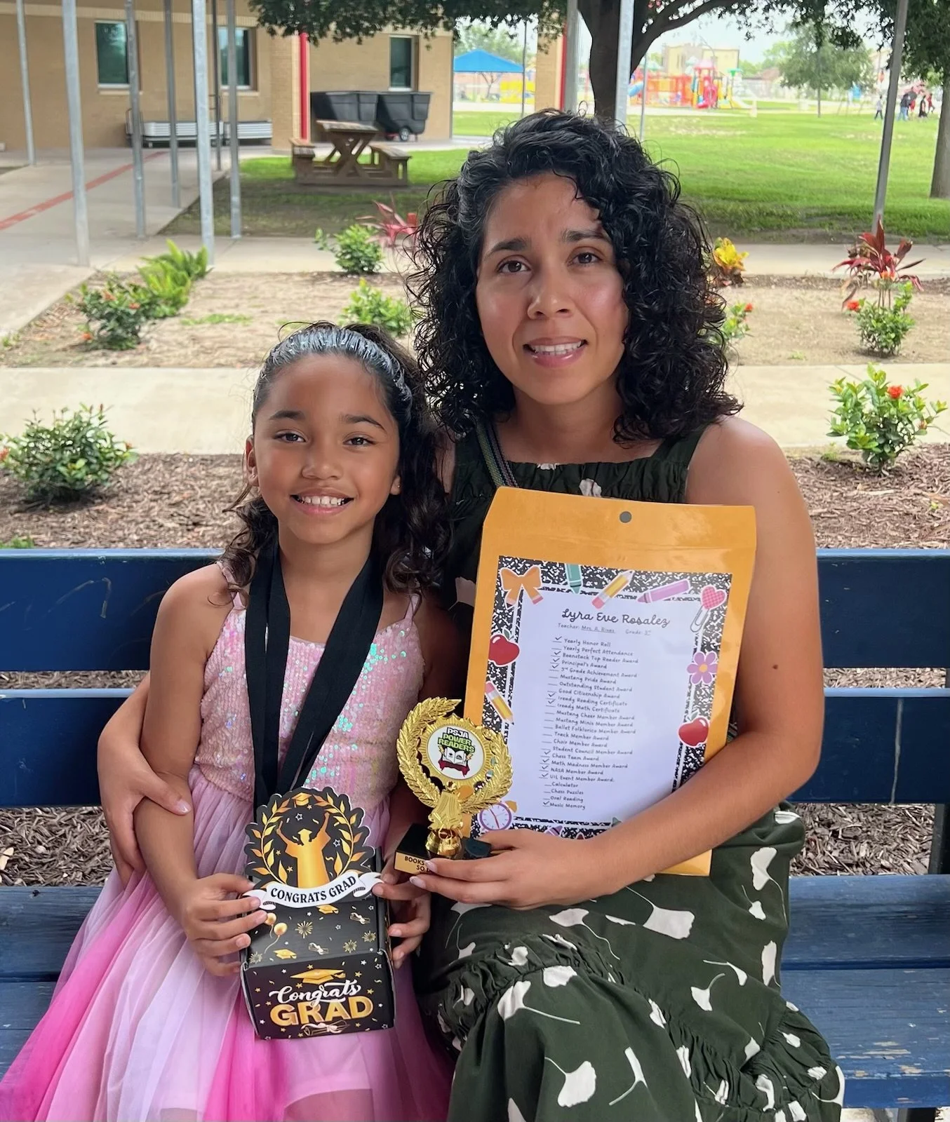 A woman and a girl sitting on a park bench celebrating a graduation. The girl is holding a 'Congrats Grad' gift bag and medals, and the woman is holding a diploma and a gold trophy. The girl is wearing a pink dress and the woman is wearing a green camo dress. The background shows a playground and park surroundings.