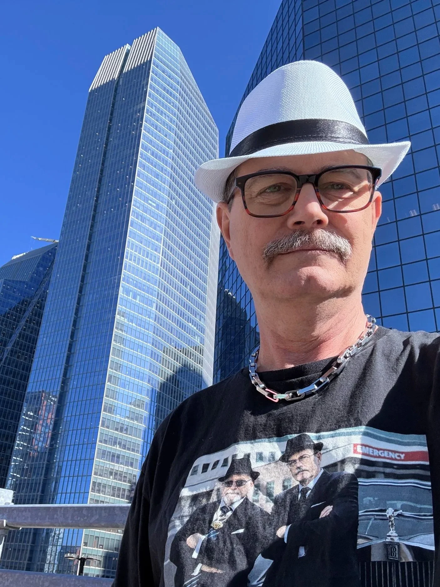 A man wearing a white trilby hat, glasses, and a chain necklace taking a selfie in front of tall blue glass office buildings on a clear sunny day.