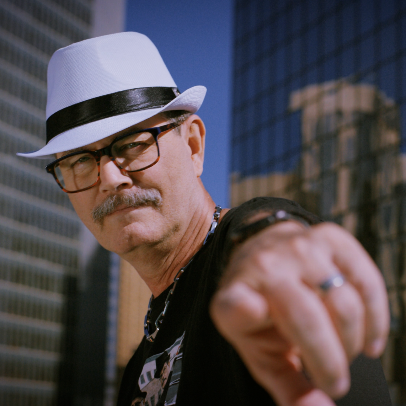 Close-up of a man wearing a white trilby hat, black-rimmed glasses, and a necklace, pointing toward the camera with a cityscape in the background.