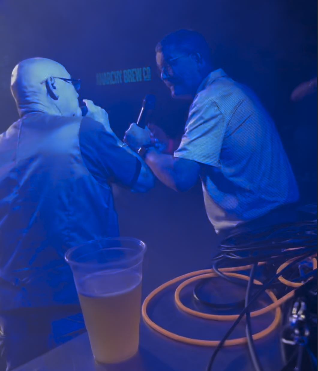 Pete (of Pete and Bas) and the artist 'story.' rapping together on stage with a sign reading 'ANARCHY BREW' visible in the background, and a plastic cup of beer on the table.