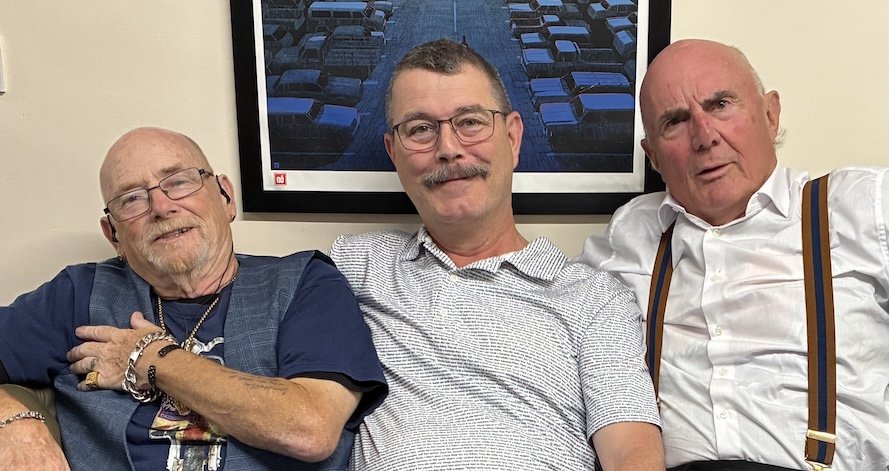 Rap artists Pete and Bas sit on either side of the artist 'story.' on a beige couch, smiling and posing for the camera, with a framed photo of a parking lot hanging on the wall behind them.