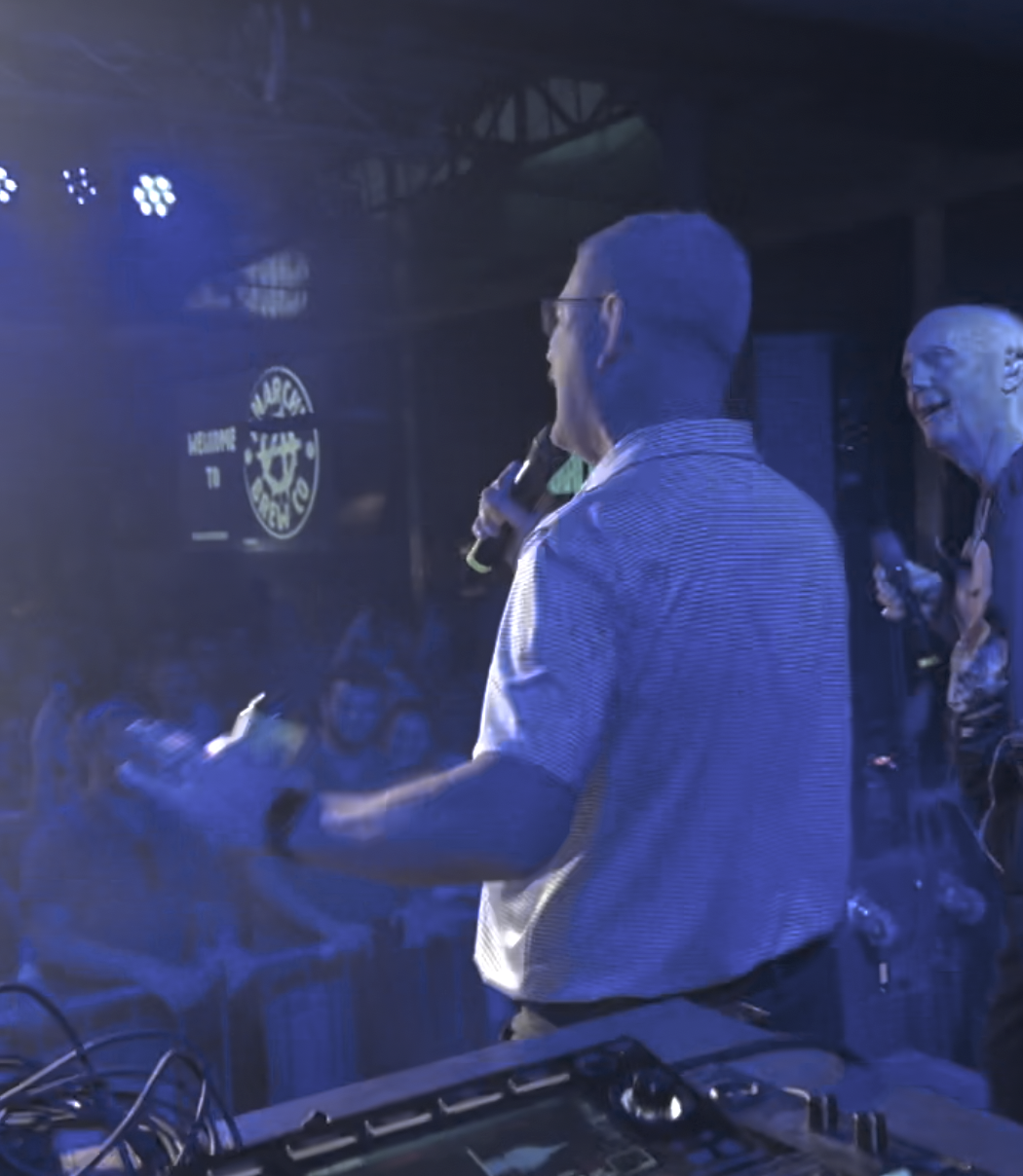 Bas (of Pete and Bas) looks surprised at the artist 'story.' on stage, rapping, with a crowd in the background at a nightclub in Newcastle, UK.