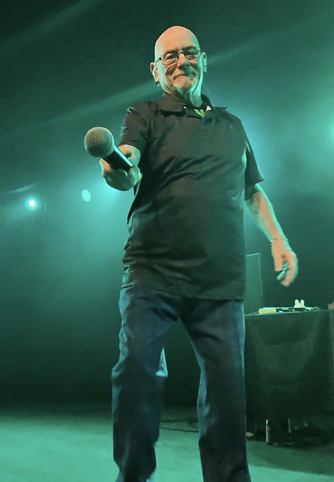 Pete (of Pete and Bas) a person with a shaved head, glasses, and earrings offers a microphone to a man in the crowd at a concert in Seattle.