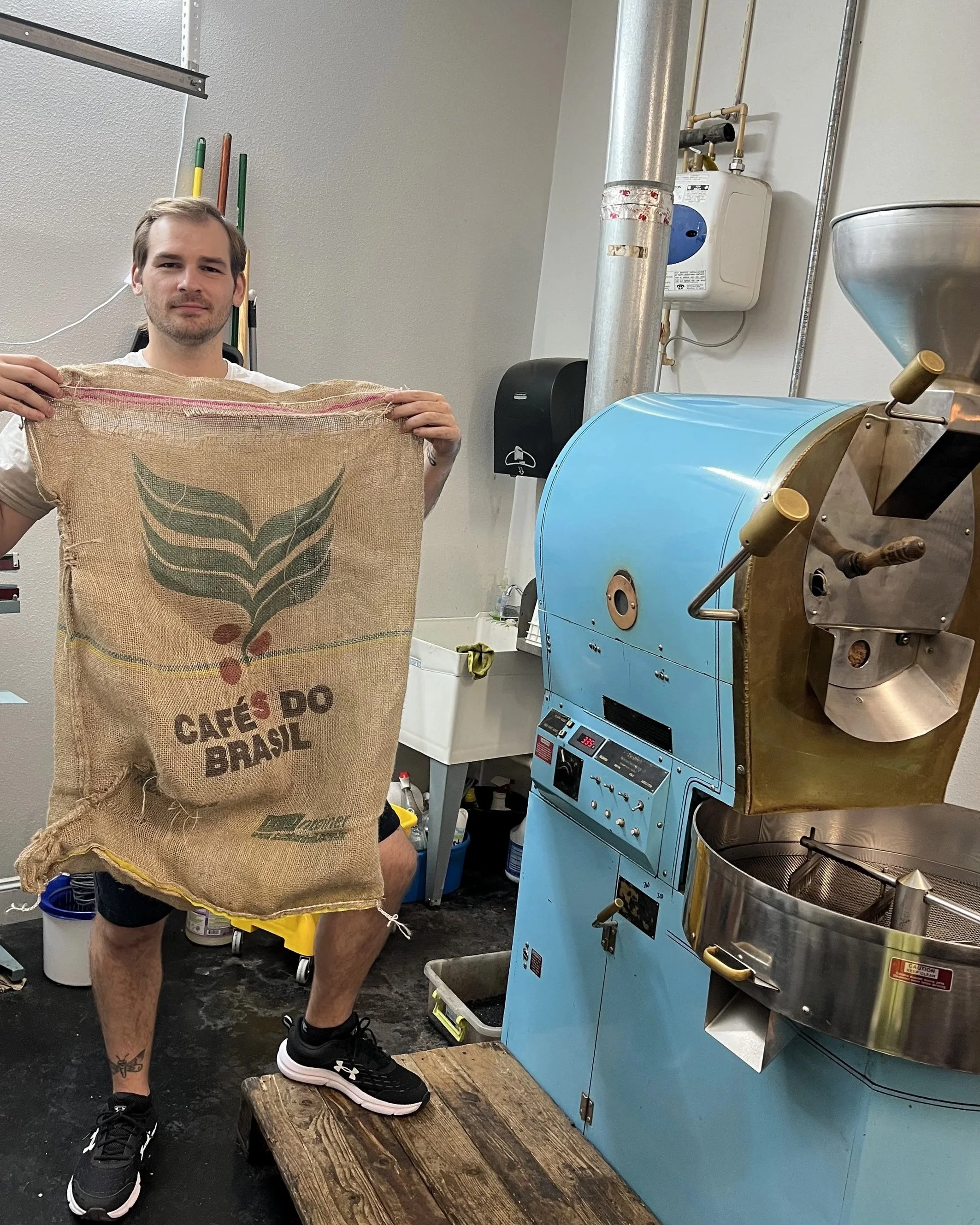 A man with tattoos on his legs wearing black sneakers and shorts stands on a wooden platform, holding a burlap bag with a green leaf logo and the words 'CAFÉS DO BRASIL,' inside a coffee roasting facility. There is a blue coffee roaster machine to his right and various tools and containers in the background.