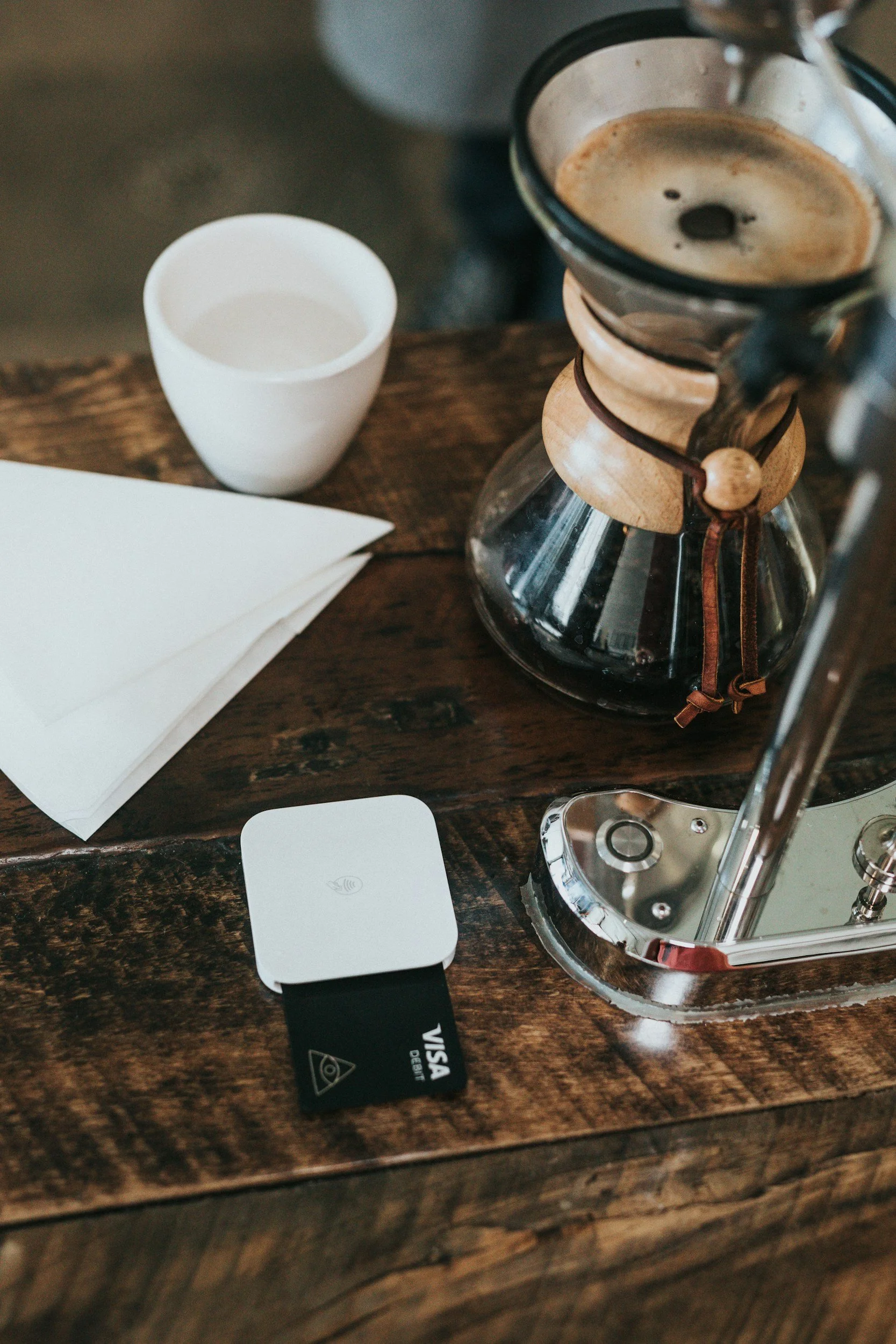 A coffee pot with brewed coffee, a white ceramic cup, a black Visa credit card, and a white smartphone are arranged on a rustic wooden table.