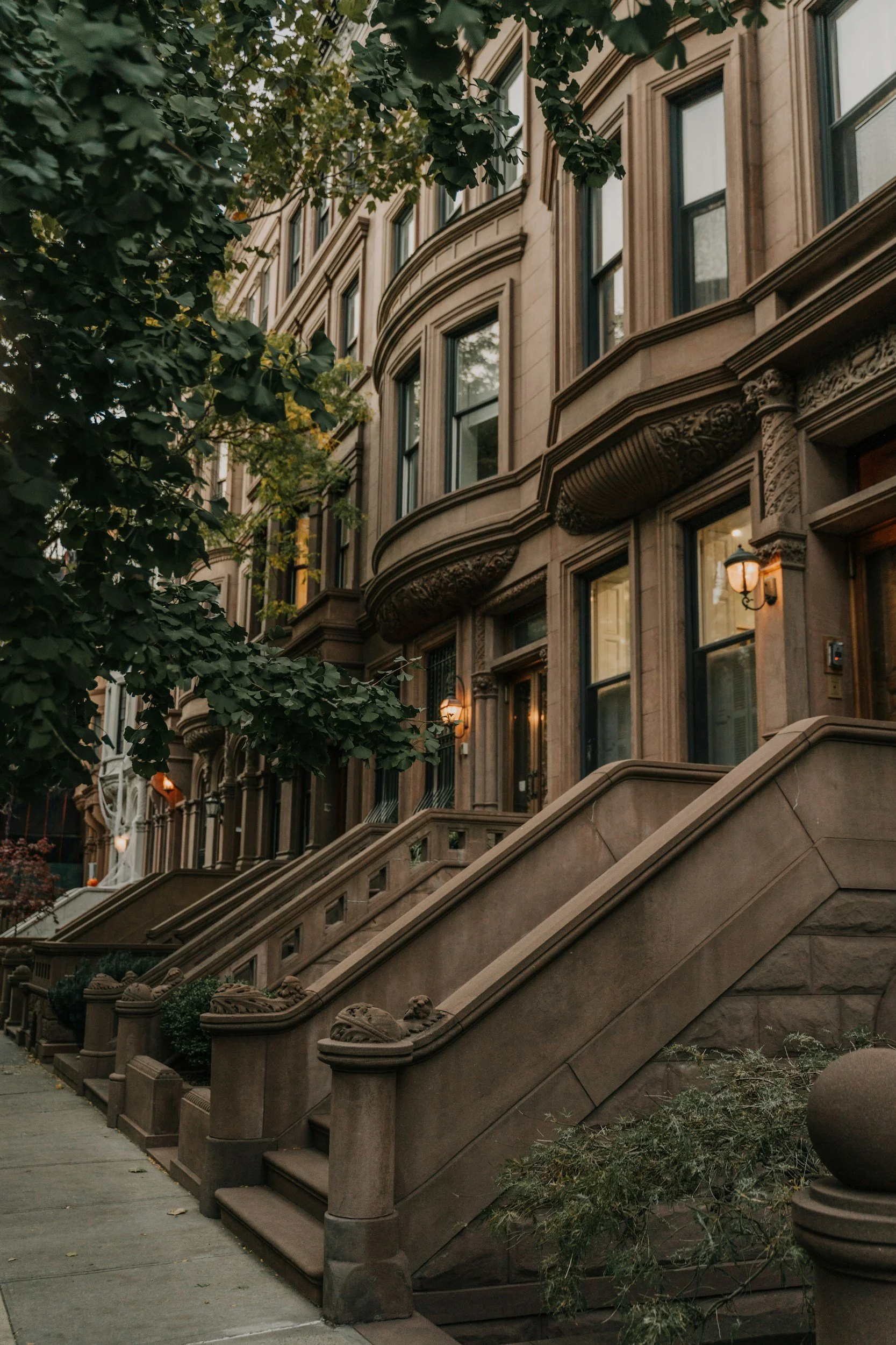 A brownstone building with detailed architectural features and stairs leading up to the entrances, with greenery in the foreground.