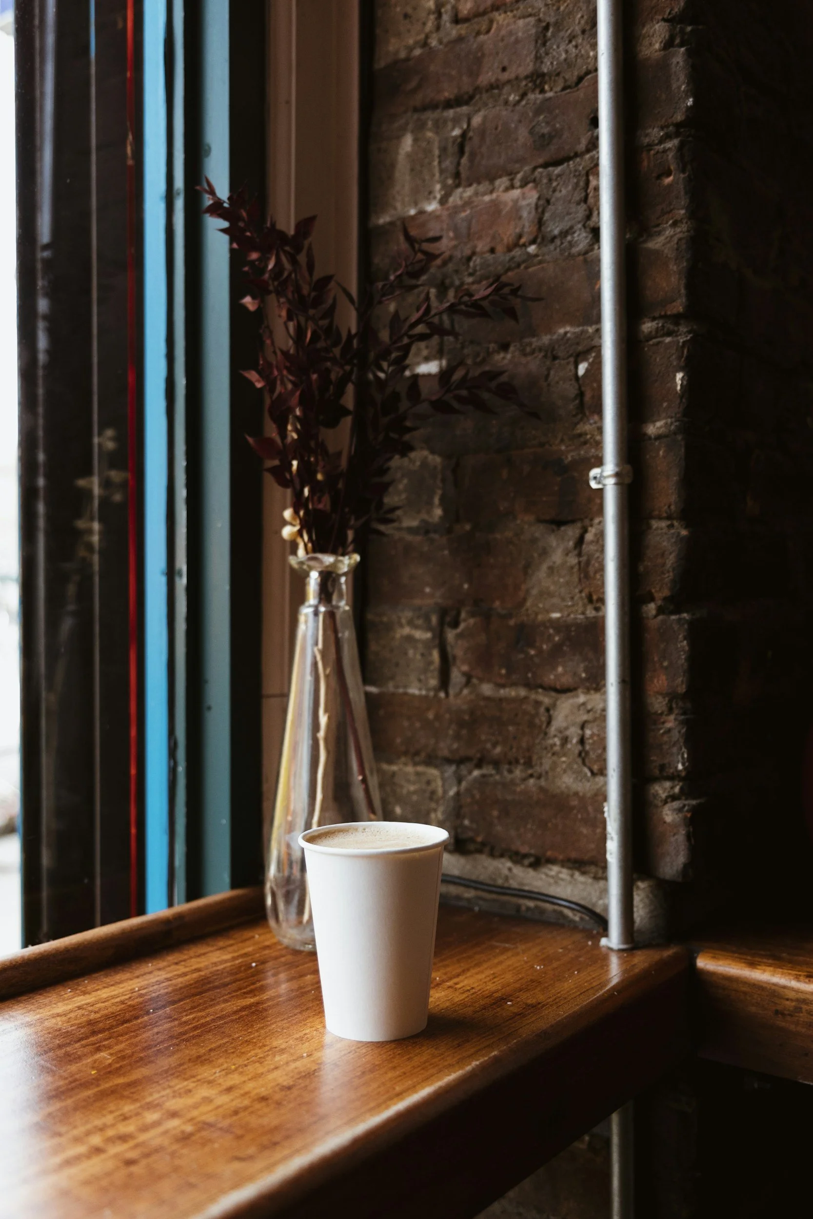 A cup of coffee on a wooden table next to a small vase with reddish-brown leaves, set against a brick wall background.